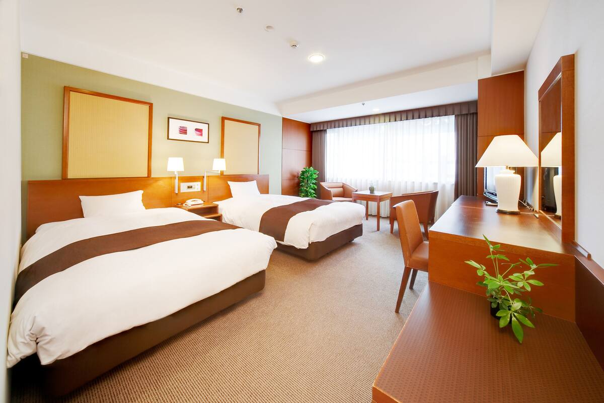 A hotel room with two beds, a desk, a chair, a small table, a plant, and a large window with curtains.