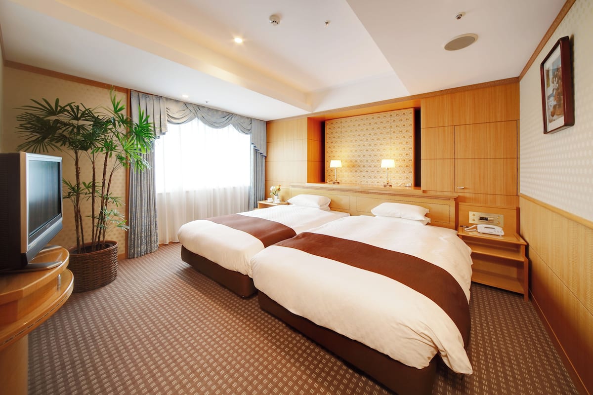 A hotel room with a large bed, a TV, a plant, and a window with curtains.