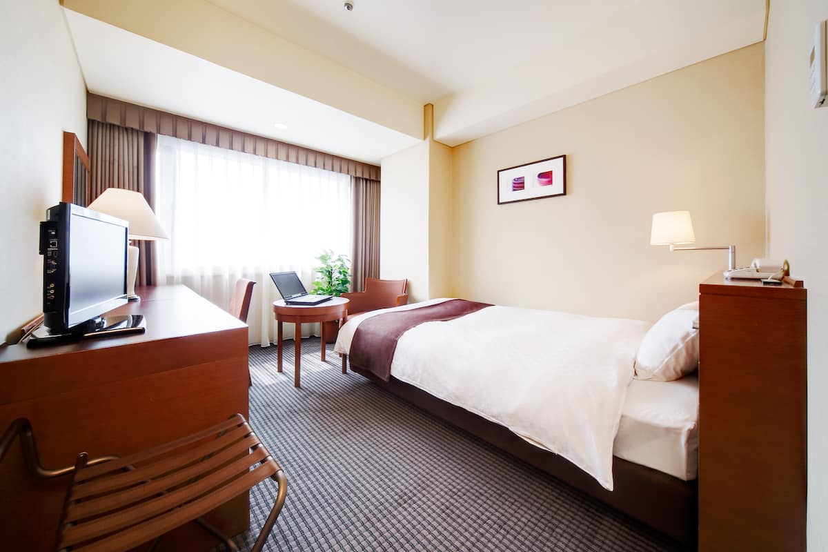 A hotel room with a bed, desk, chair, and a desk lamp.