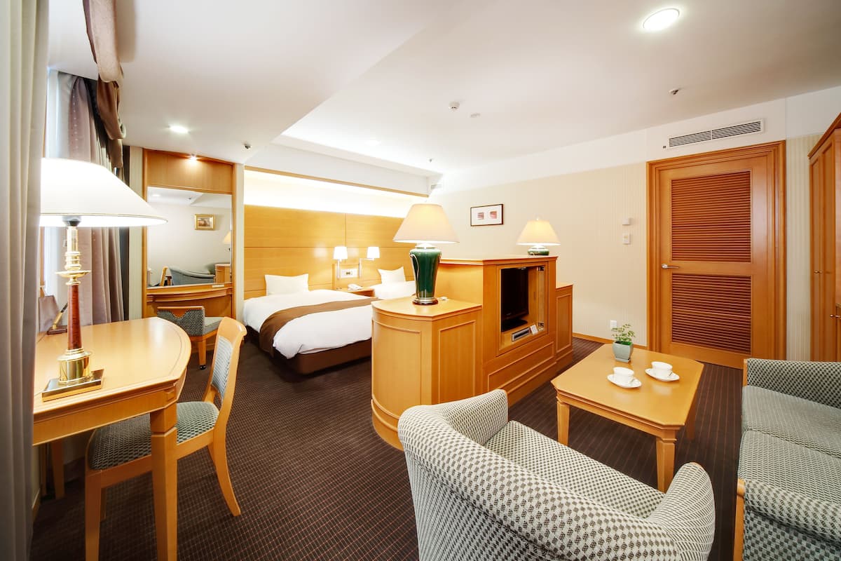 A hotel room with a bed, a desk with a lamp, a TV, and a seating area with chairs.