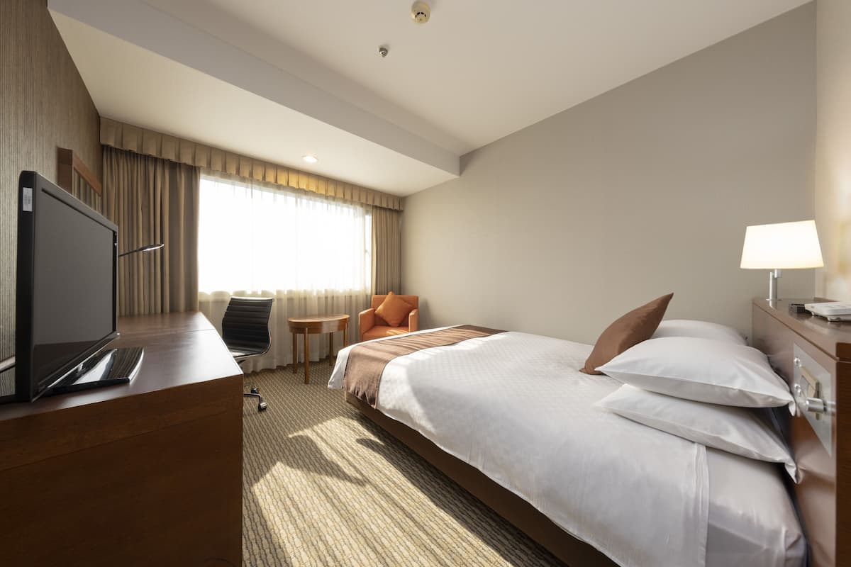 A hotel room with a bed, a desk with a computer, a chair, a lamp, and a window with curtains.