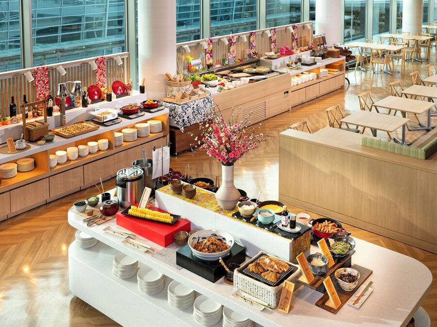 A buffet setup with various food items and utensils, a vase with flowers, and a seating area with tables and chairs.