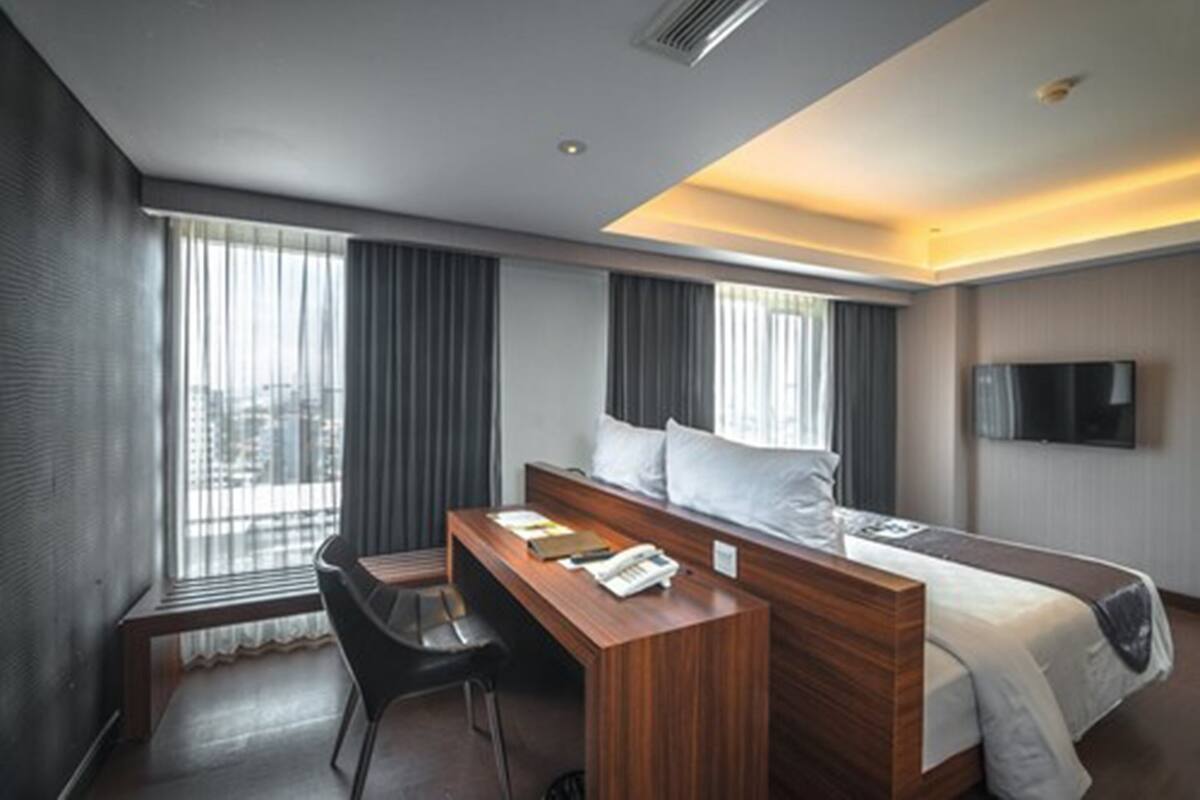 A hotel room with a bed, a desk, and a chair. There is a window with curtains, a television on the wall, and a city view.