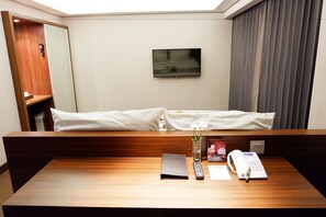 Une chambre d’hôtel avec un bureau en bois, un téléviseur à écran plat fixé au mur, un lit avec une literie blanche et une grande fenêtre avec des rideaux.