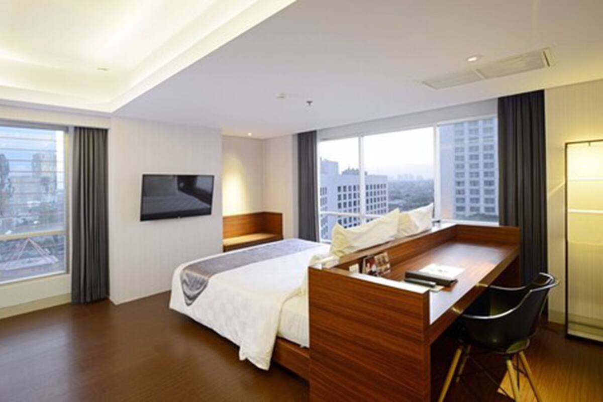 A modern hotel room with a large bed, a desk, and a chair. There is a TV mounted on the wall and a window with a city view.