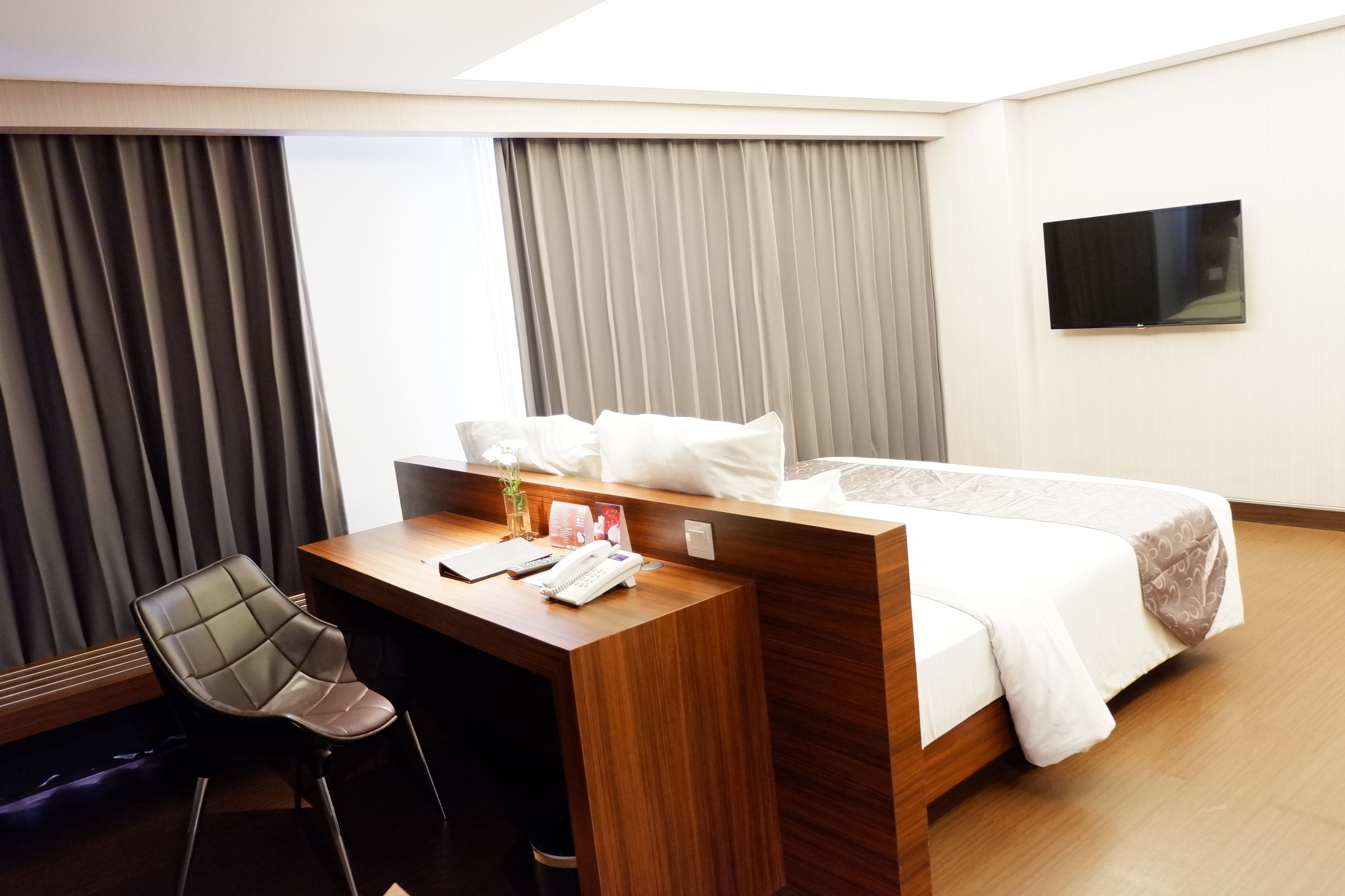 A hotel room with a wooden desk, a chair, a bed with white bedding, and a flat-screen TV mounted on the wall.