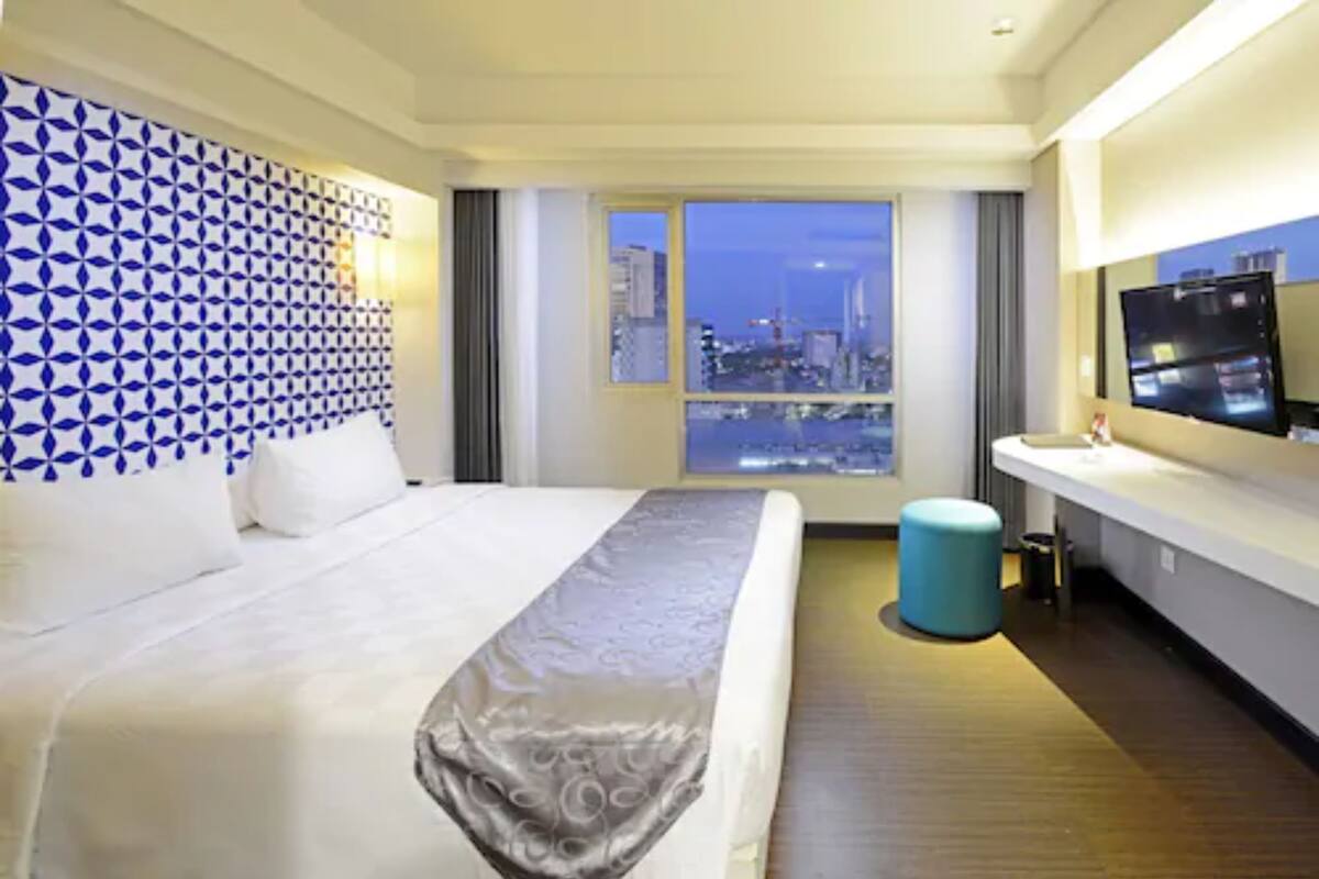A hotel room with a large bed, a view of a cityscape through a window, a blue patterned wall, and a modern bathroom with a sink and mirror.