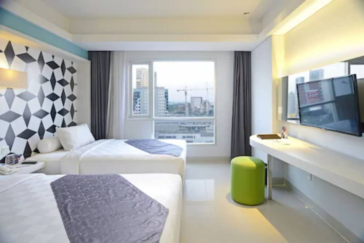 A hotel room with two beds, a large window with a city view, a flat-screen TV, and a green ottoman.