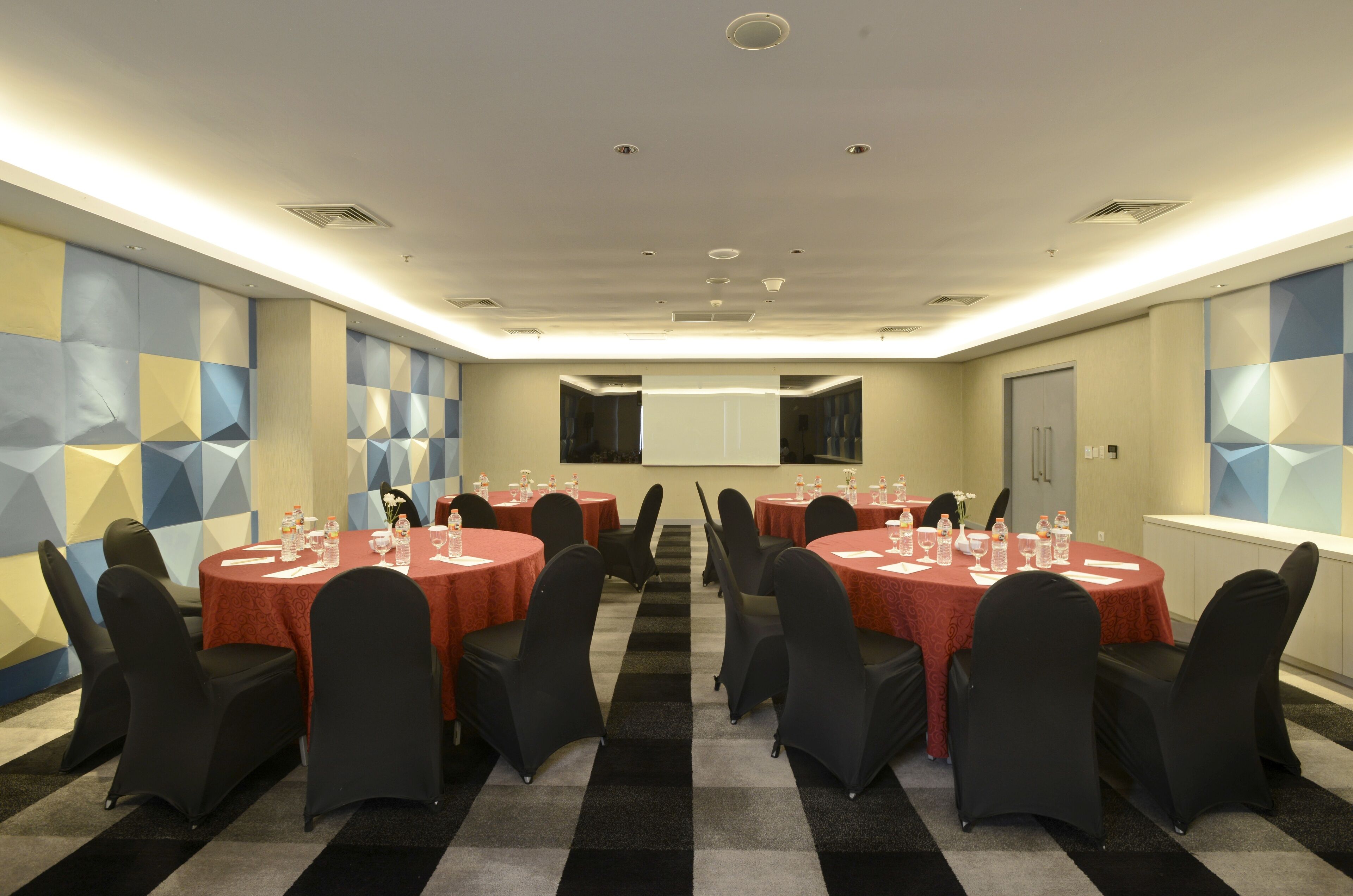 Une salle de conférence dotée de plusieurs tables et chaises, équipée de bouteilles d’eau et de verres, et un écran de projection fixé au mur.