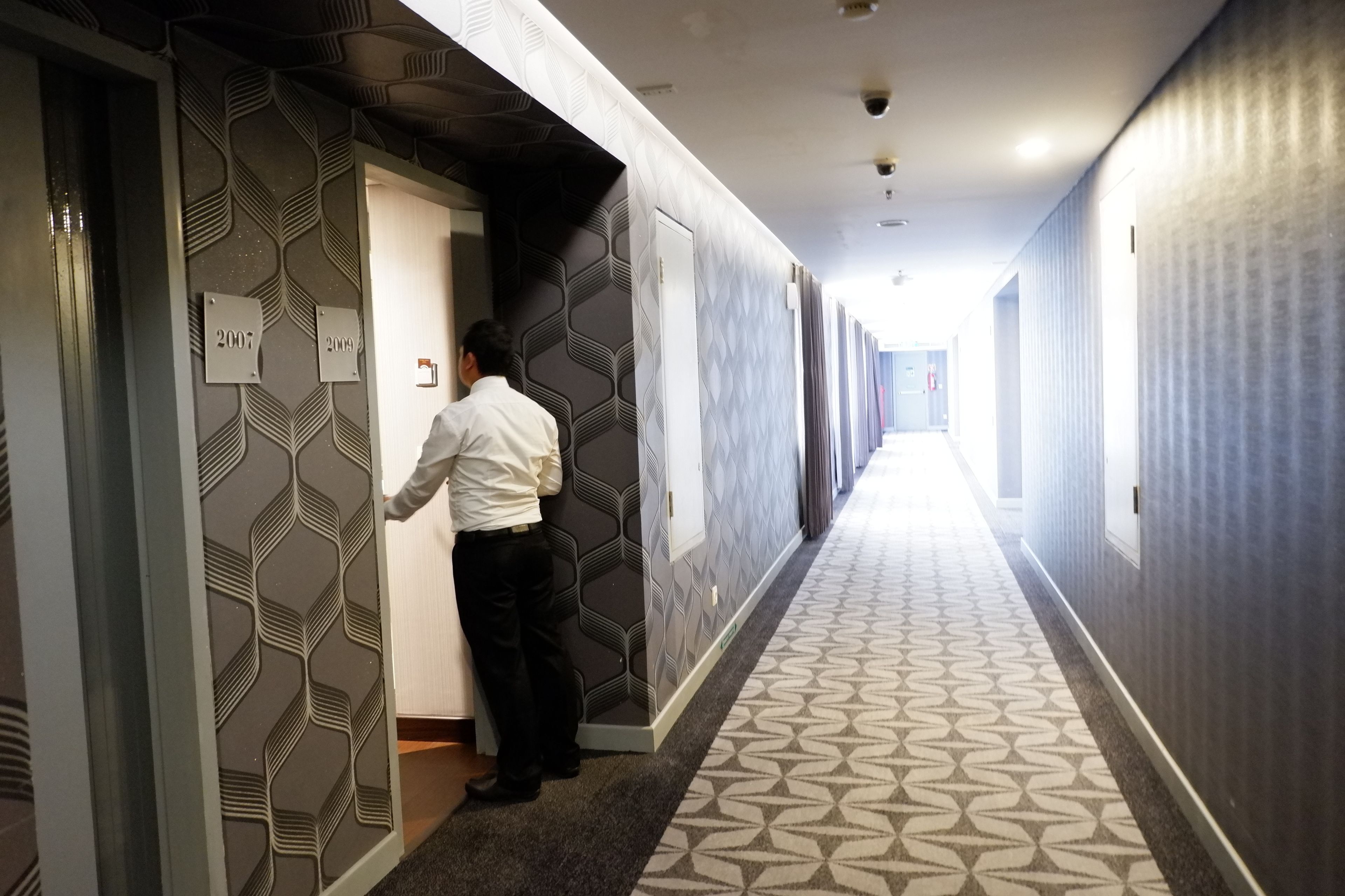 A hotel corridor with patterned walls and carpet, a man at an elevator, and doors marked with years.
