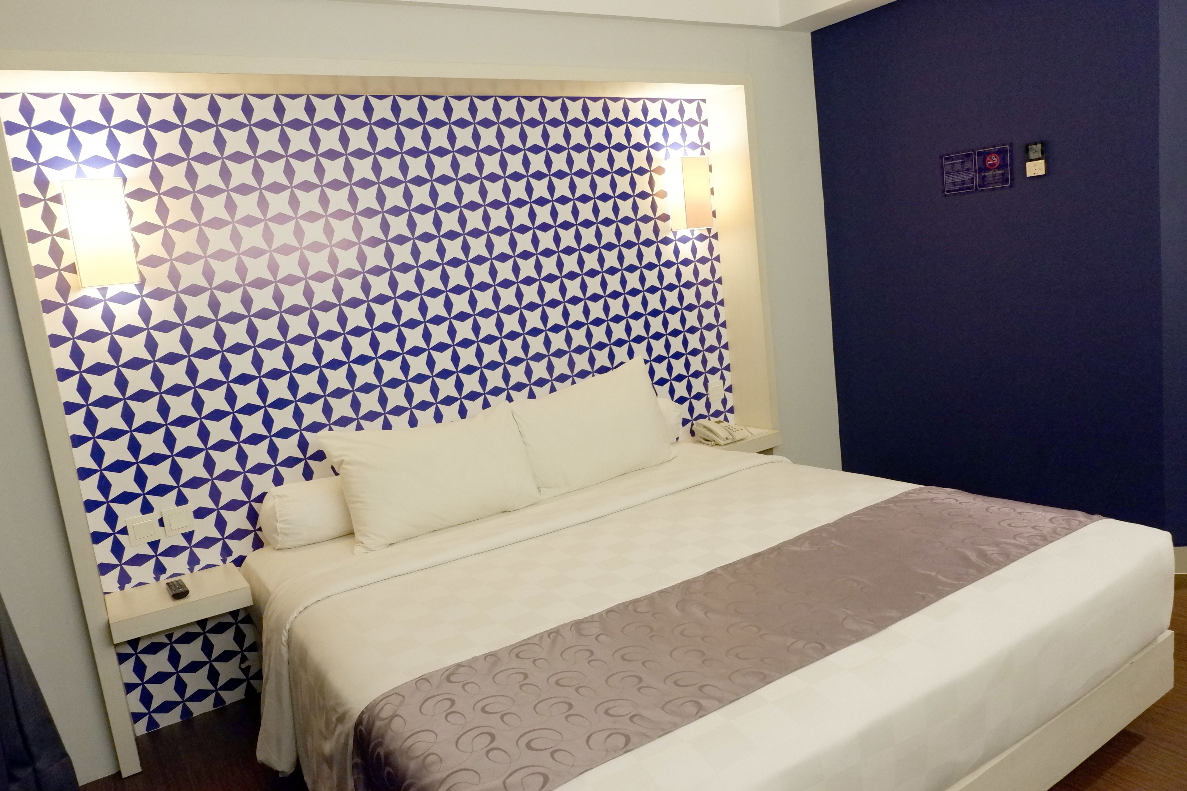 Uma cama bem arrumada com cabeceira estampada, uma mesa de cabeceira com controle remoto e uma parede com padrão geométrico.