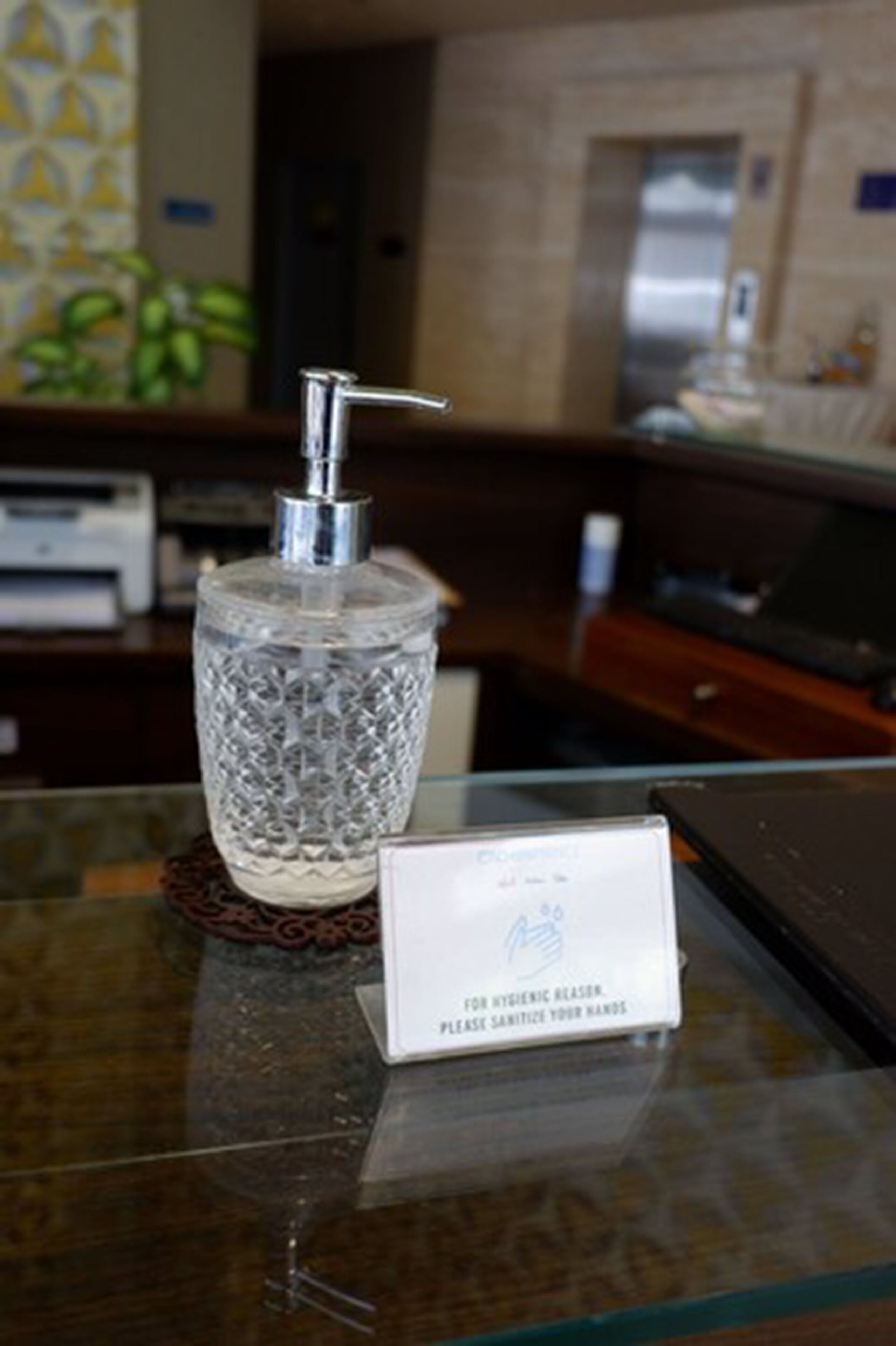 A glass dispenser with a pump, a sign instructing to sanitize hands, and a blurred background with a plant and a door.