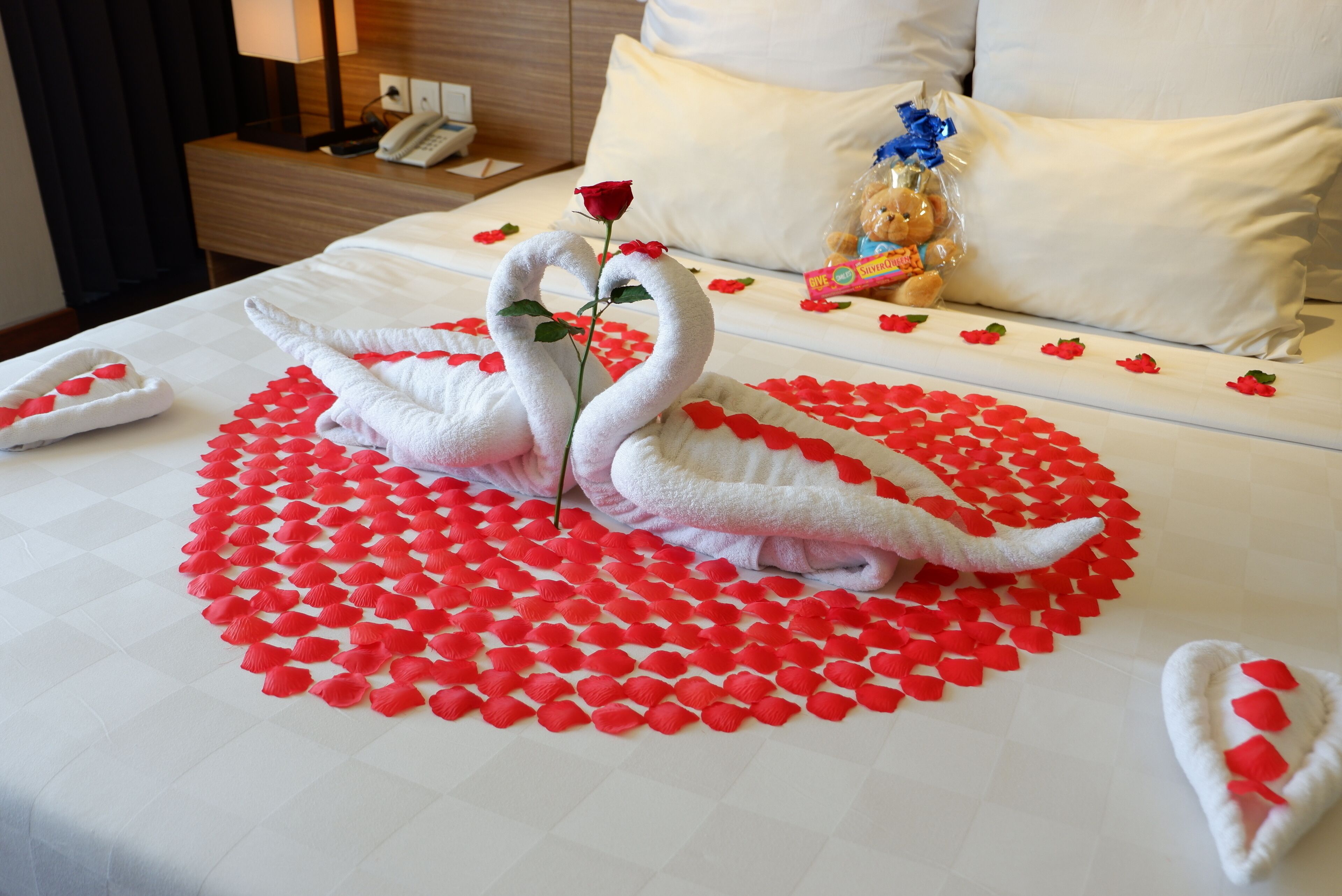 A neatly made bed with a heart-shaped arrangement of red rose petals, two swan-shaped towels, and a single rose.