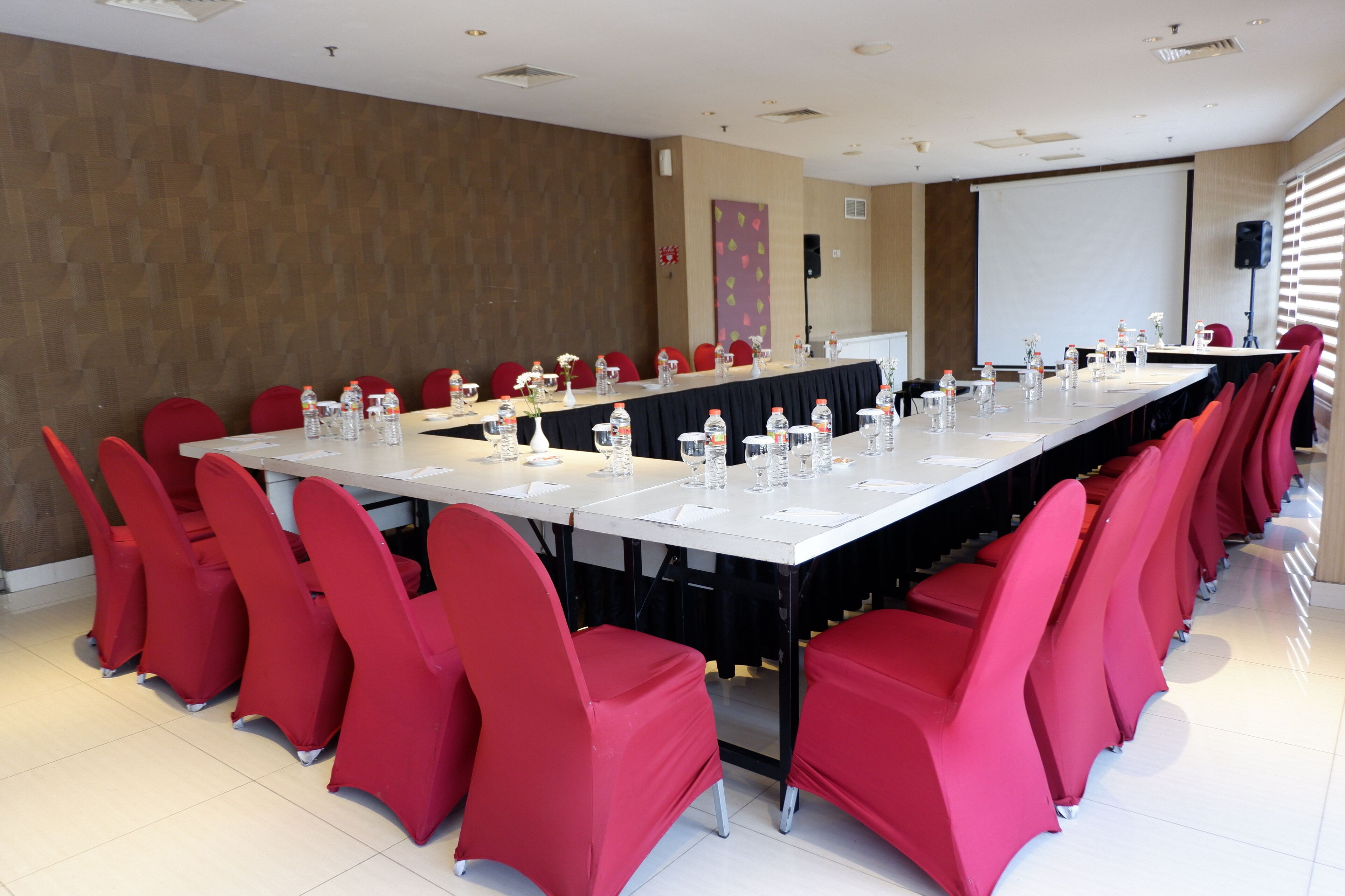 Un salón de conferencias con una mesa larga, sillas rojas, botellas de agua y una pantalla de proyector.