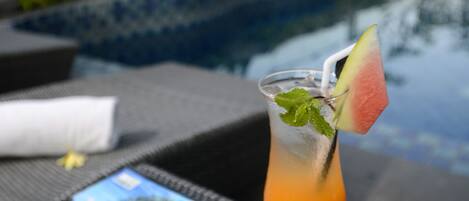 Un verre à cocktail, une tranche de pastèque et quelques feuilles de menthe, le tout posé sur une table au bord d’une piscine.