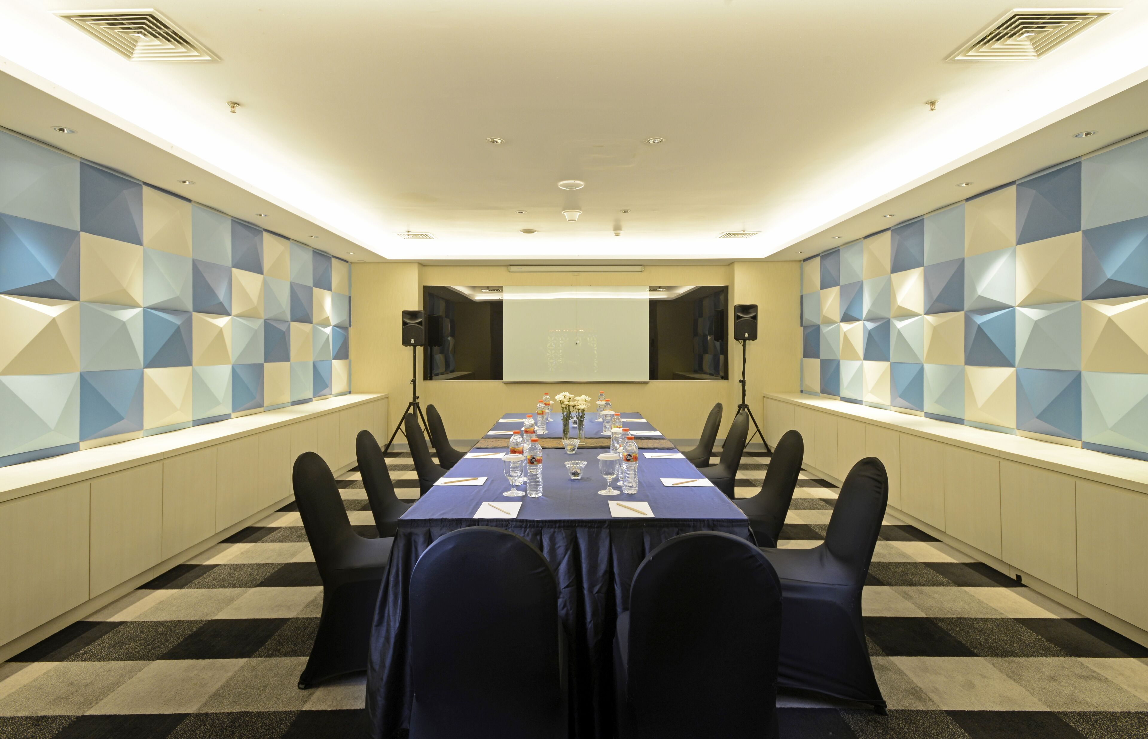 A conference room with a long table, chairs, a projector screen, and geometric wall panels.