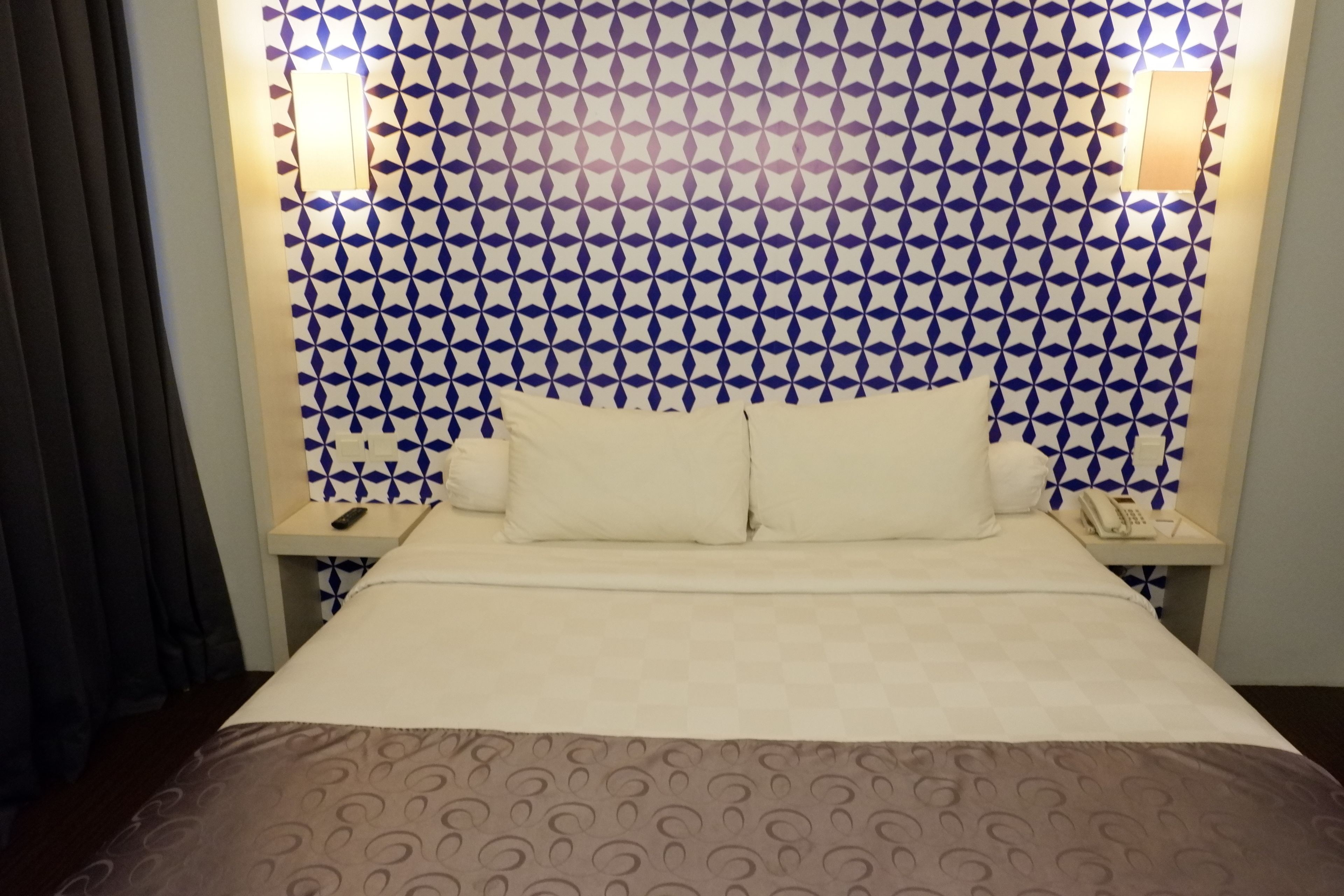 A hotel room with a patterned wallpaper, a bed with two pillows, a bedside table with a phone, and a nightstand with a lamp.