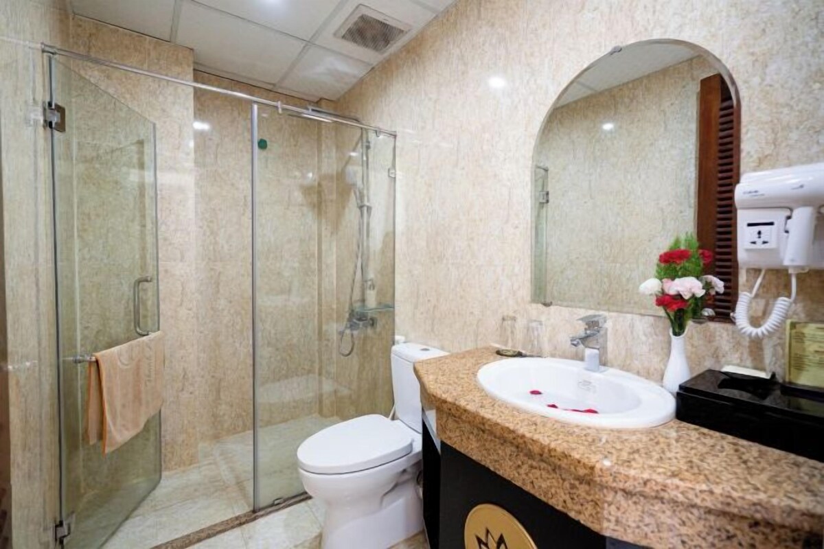 Une salle de bain avec une douche à parois de verre, des toilettes blanches, un lavabo surmonté d’un plan de travail en granit et un miroir agrémenté d’un vase à fleurs.