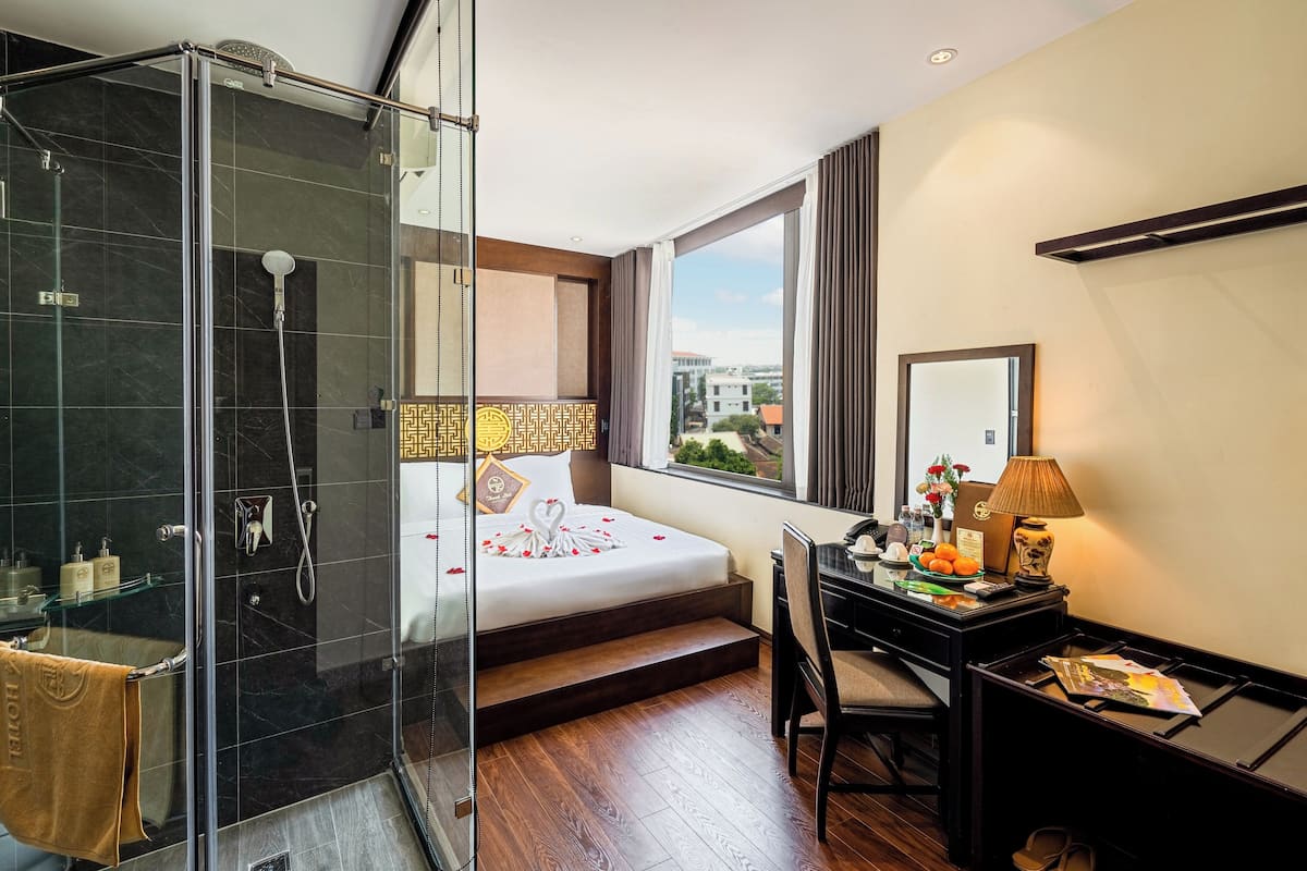 Une chambre d’hôtel avec une douche en verre, un lit parsemé de pétales de rose, un bureau avec une lampe et une vue sur les bâtiments par la fenêtre.