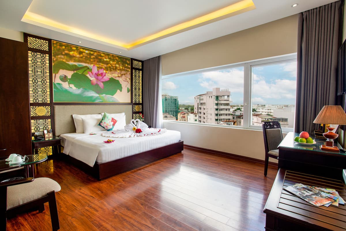Une chambre d’hôtel avec un grand lit, un bureau avec une lampe, une chaise et une vue sur la ville par une grande fenêtre.