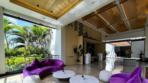 A spacious indoor area with a modern design, featuring a purple sofa, round coffee tables, and a view of lush greenery outside. - ILLIRA Lite Praya Lombok (Praya)