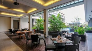 A modern restaurant with wooden flooring, black chairs, and tables. It has large windows offering a view of a pool and greenery. - ILLIRA Lite Praya Lombok (Praya)