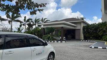 Un auto blanco estacionado frente a un edificio con entrada techada, palmeras y cielo despejado.