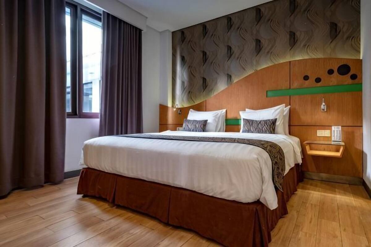 A hotel room with a large bed, wooden flooring, a window with curtains, and a decorative wall panel.