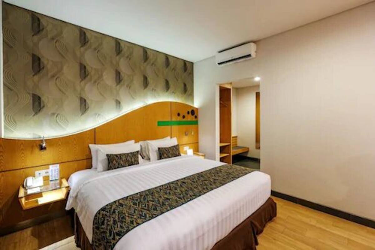A hotel room with a large bed, a wooden headboard, a nightstand, and a wall-mounted air conditioning unit.