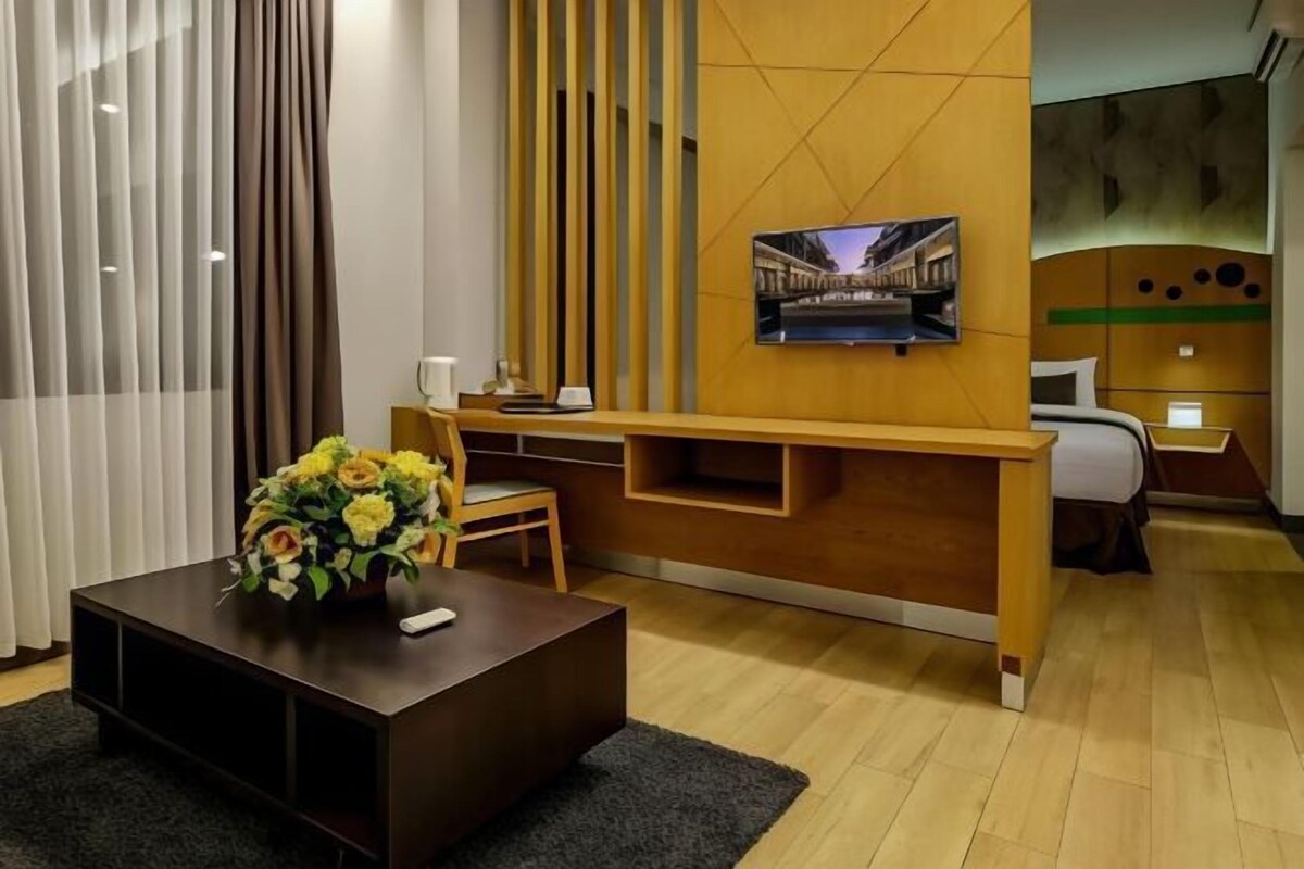 A modern hotel room with a wooden desk, a flat-screen TV mounted on the wall, a bed with a green headboard, and a small seating area with a chair and a table.