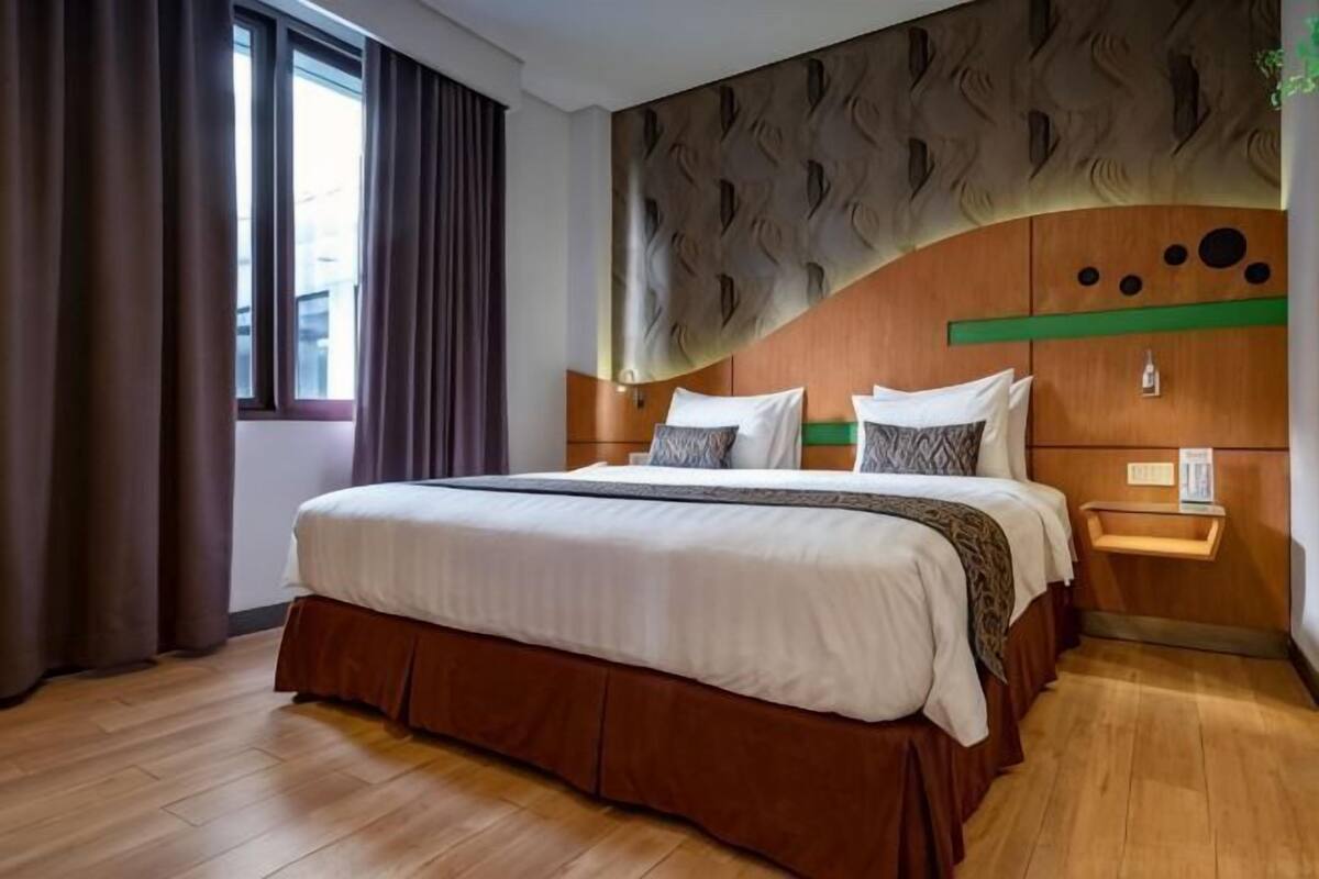 A hotel room with a large bed, wooden flooring, a headboard with a unique design, a bedside table, and a window with curtains.