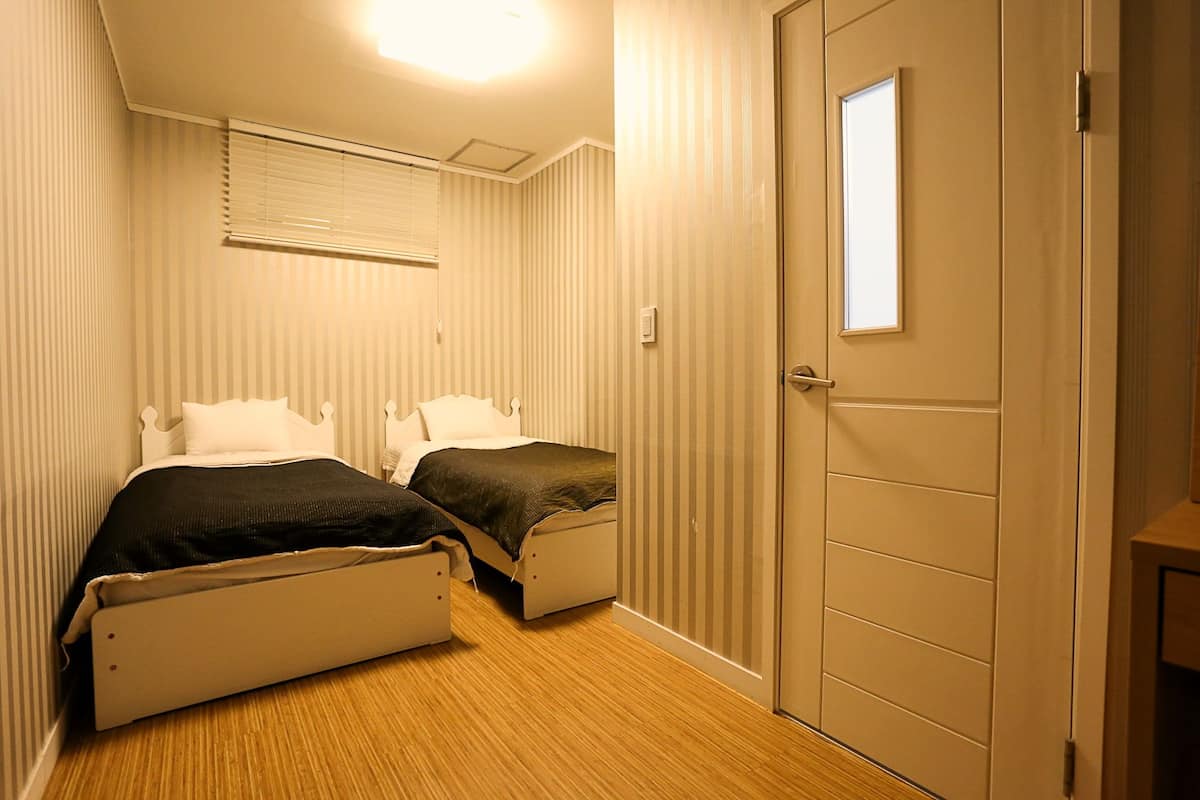 A small, narrow room with two single beds, a window with blinds, and a door with a small window.