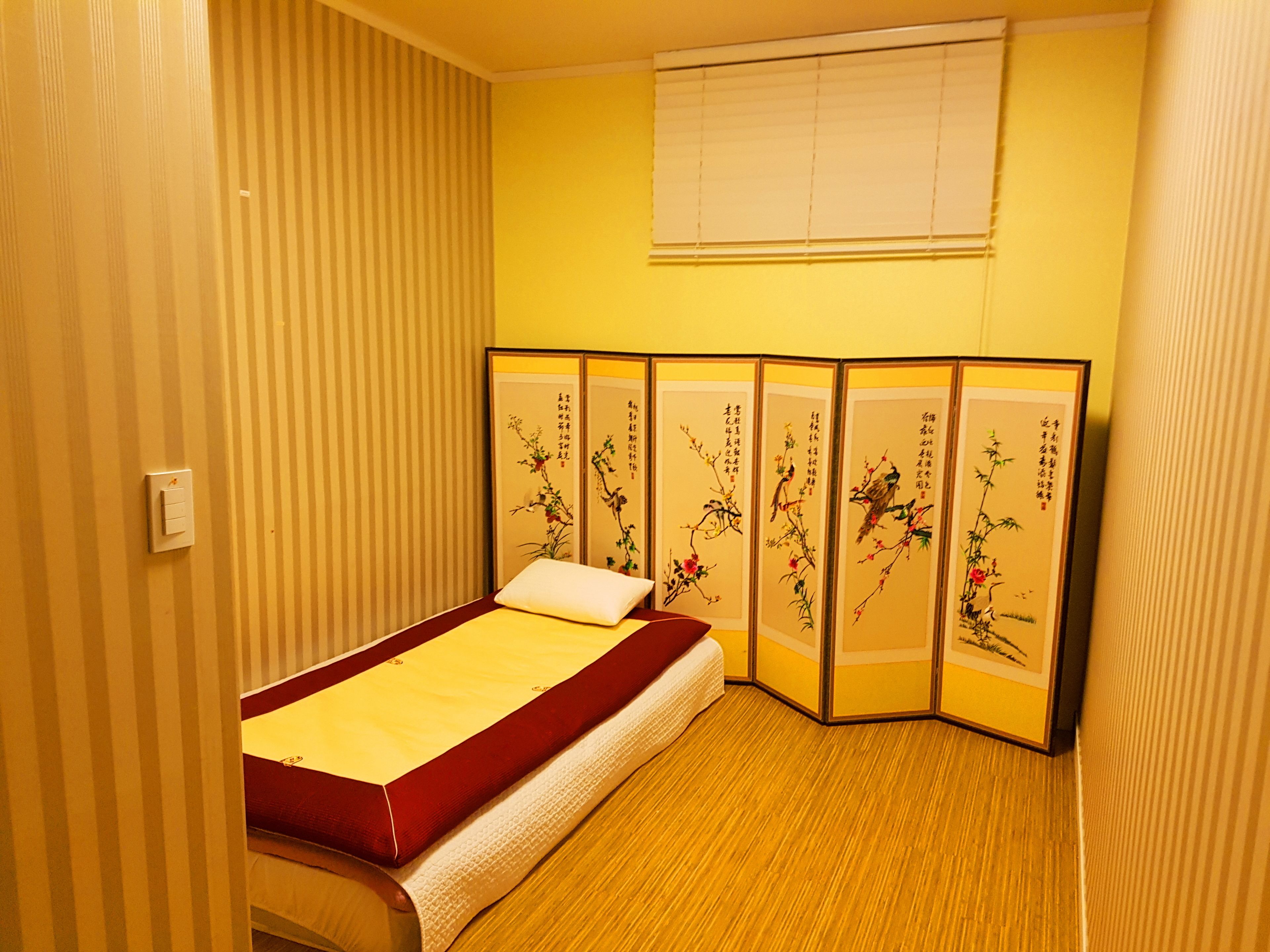 A room with a bed, a sliding shoji screen with artwork, a window with blinds, and a light switch on the wall.