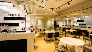 A modern restaurant with wooden flooring, white tables, and black chairs. There are multiple dining areas and a kitchen visible in the background.