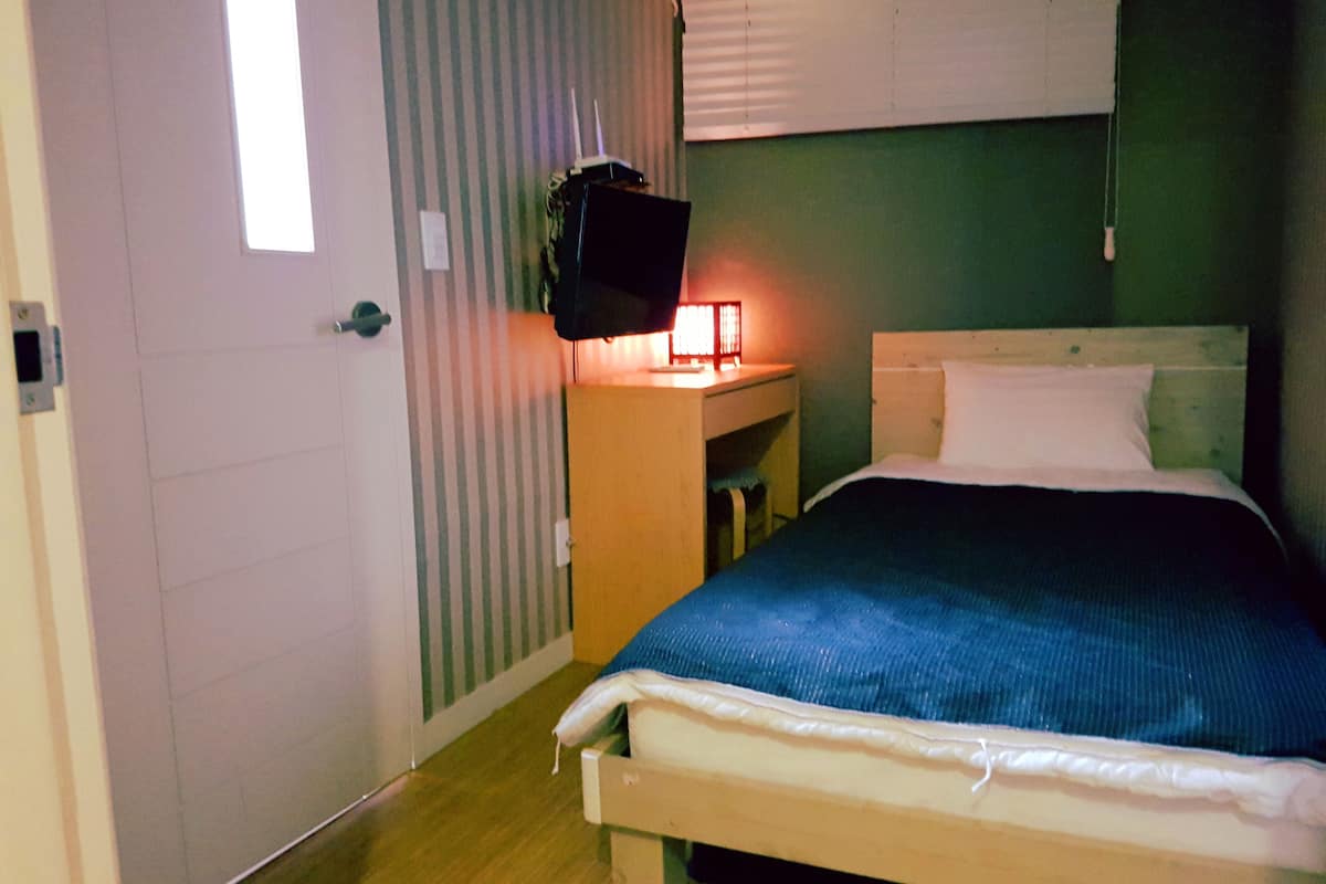 A bedroom with a bed, a bedside table with a lamp, a television on a stand, and a window with blinds.
