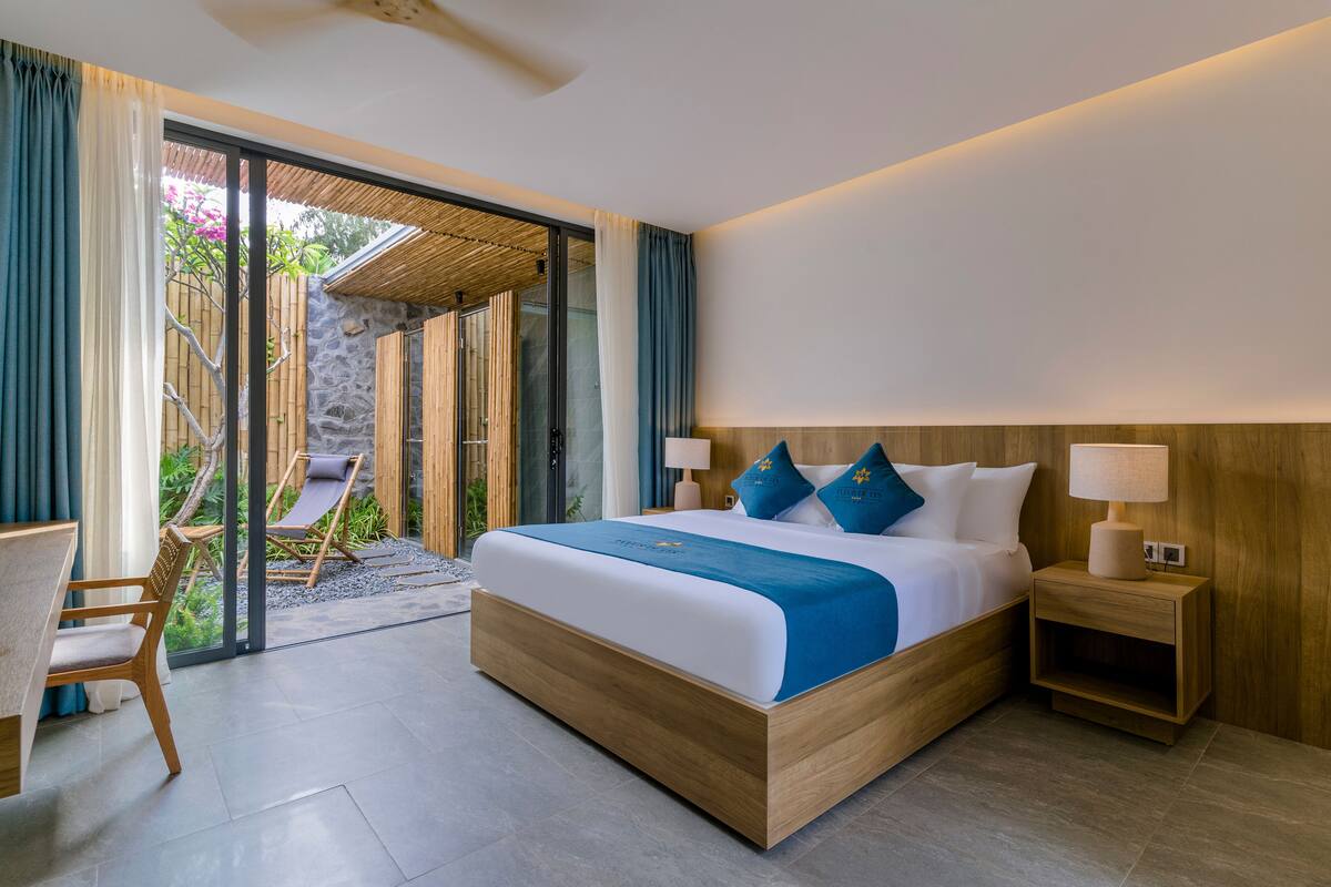 Une chambre d’hôtel moderne dotée d’un grand lit, de tables de chevet en bois, d’un coin salon avec une chaise et une table, et offrant une vue sur l’extérieur grâce à une porte vitrée.