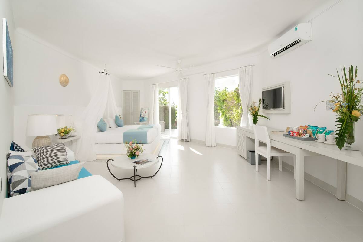 Une chambre d’hôtel moderne, décorée dans des tons de blanc, comprenant un lit, un coin salon avec une table basse, un bureau avec une chaise et un téléviseur.
