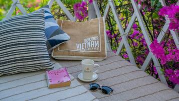 A balcony with a view of green hills, a white railing, and a table with a striped pillow, a book, a cup, and sunglasses.