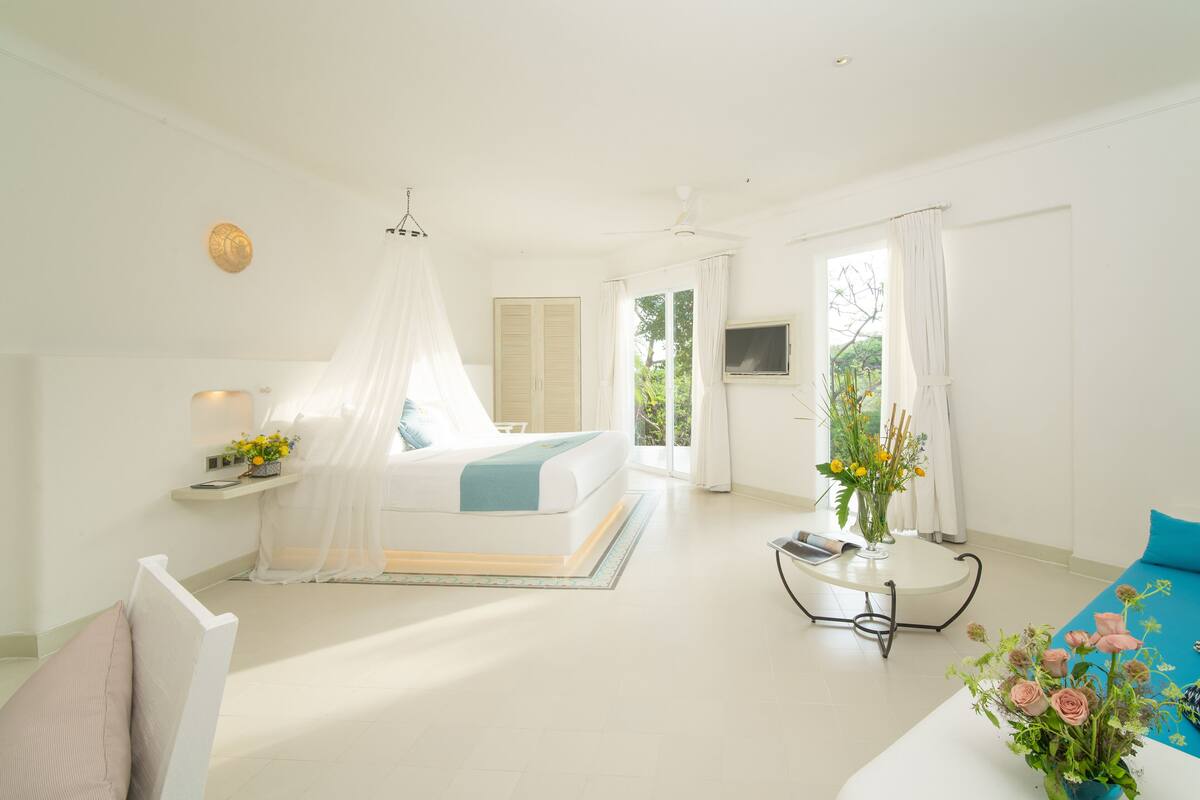 Une chambre à coucher lumineuse et spacieuse, dotée d’un grand lit, d’une table de chevet, d’une petite table ornée d’un vase de fleurs et offrant une vue sur la verdure environnante.