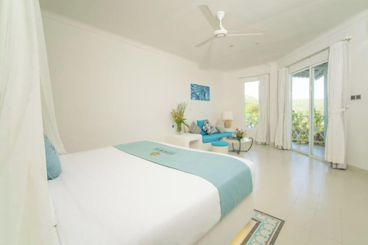 A bedroom with a bed, a ceiling fan, a window with curtains, and a view of greenery outside.