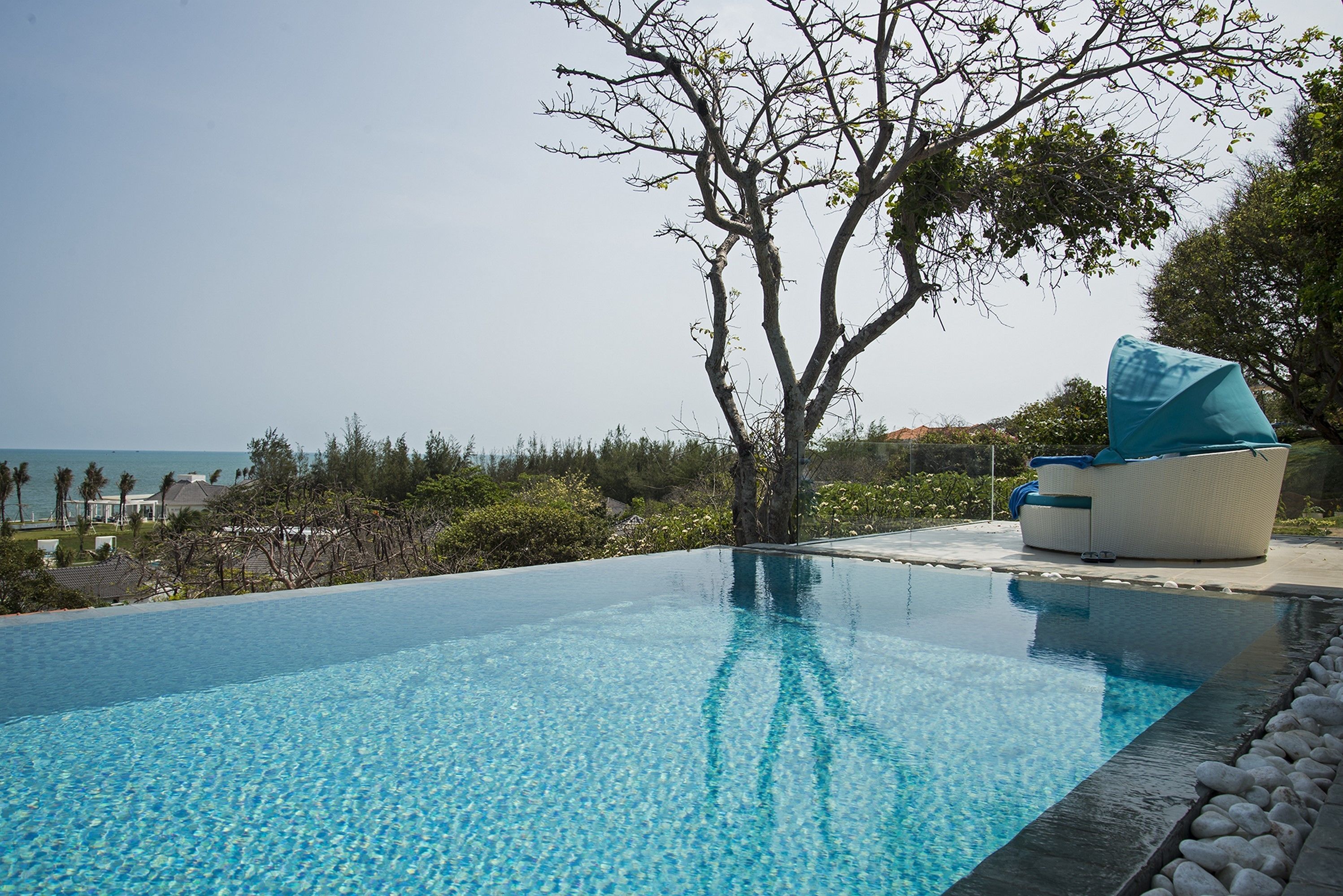 Ein Pool ohne erkennbare Kante mit Blick auf das Meer, umgeben von üppiger Vegetation und einem weißen Liegestuhl mit blauer Auflage.