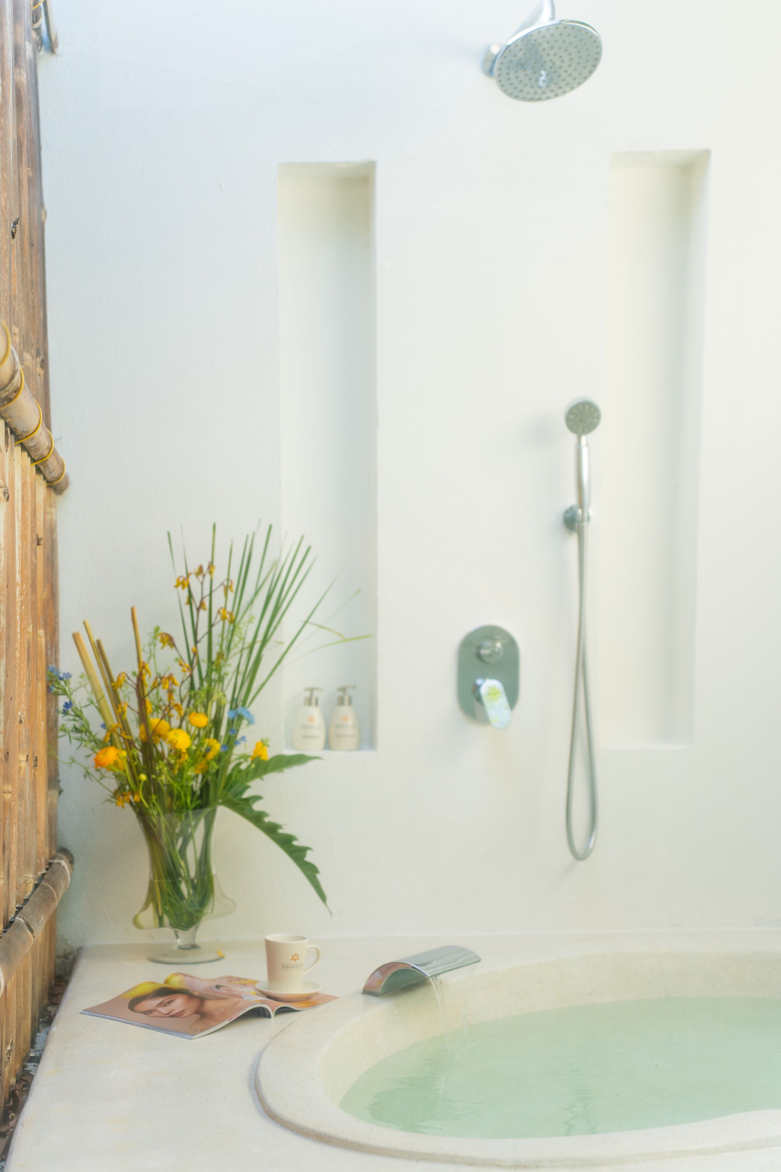 Une salle de bain avec une baignoire, une douche, une porte en bois, un vase avec des fleurs et un magazine posé sur le sol.