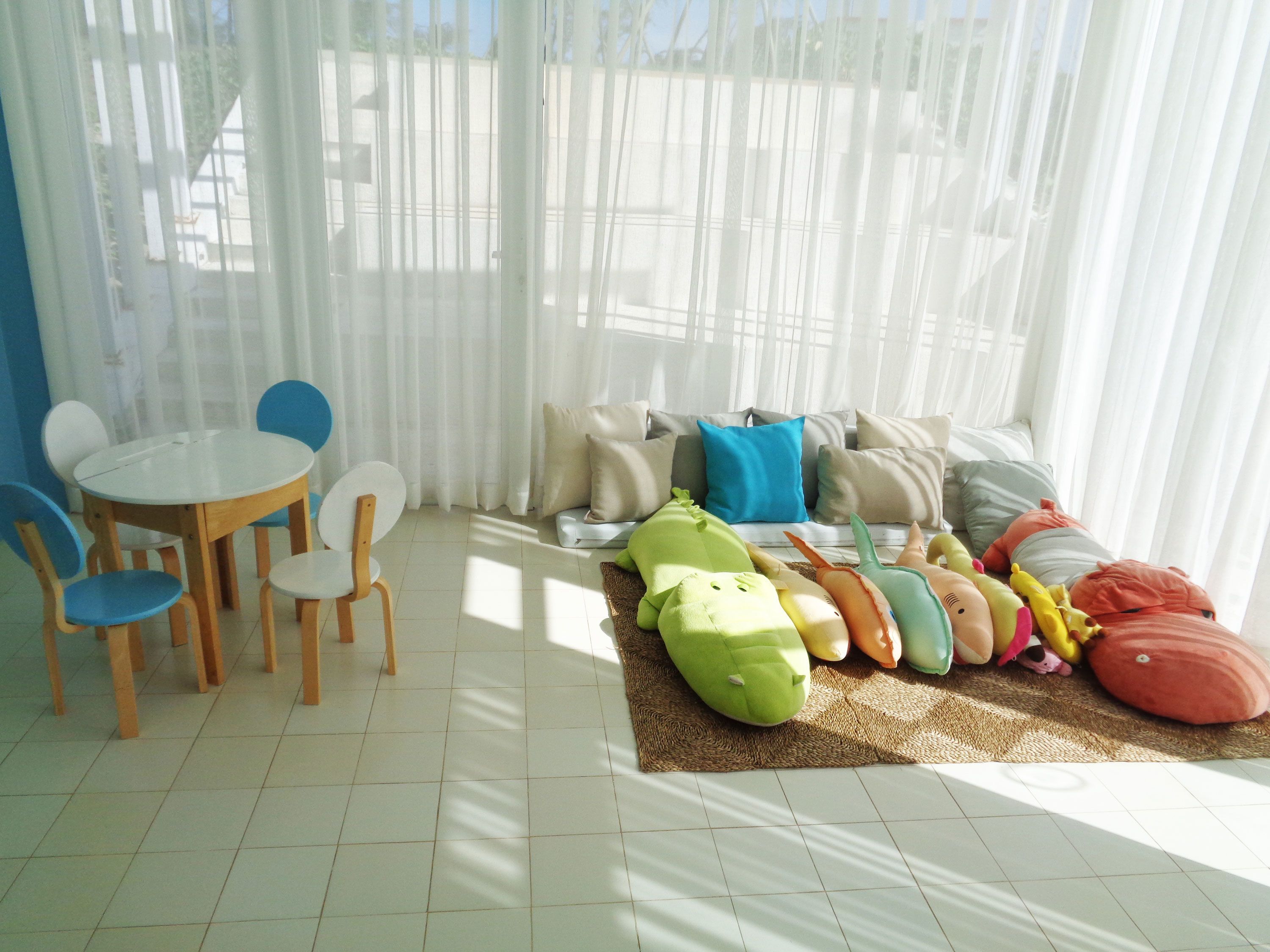A bright room with a round table and chairs, a sofa with cushions, and a rug with plush toys.