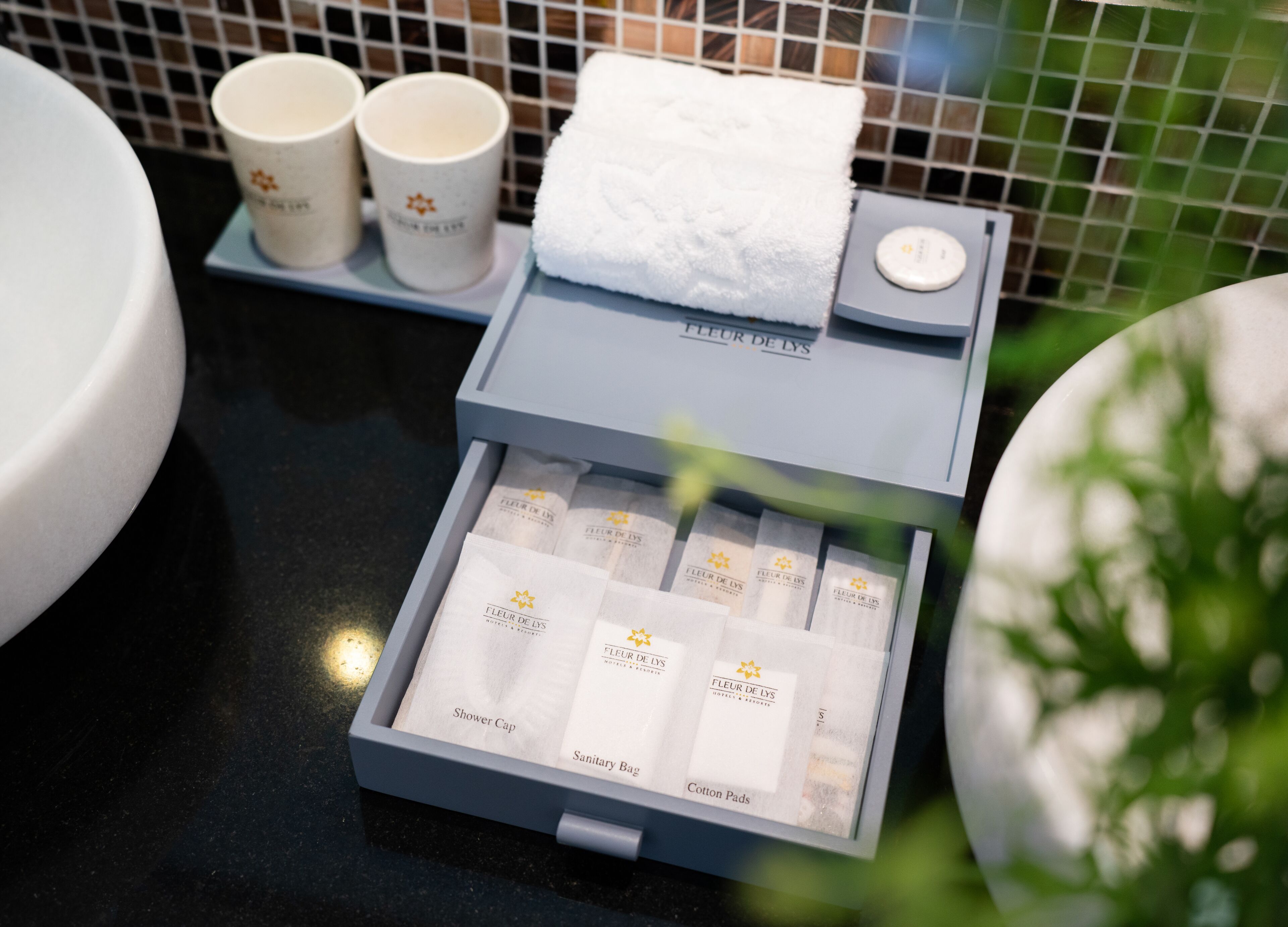 A bathroom with a white sink, two white cups, a white towel, and a box of toiletries including a shower cap, sanitary bag, and cotton pads.