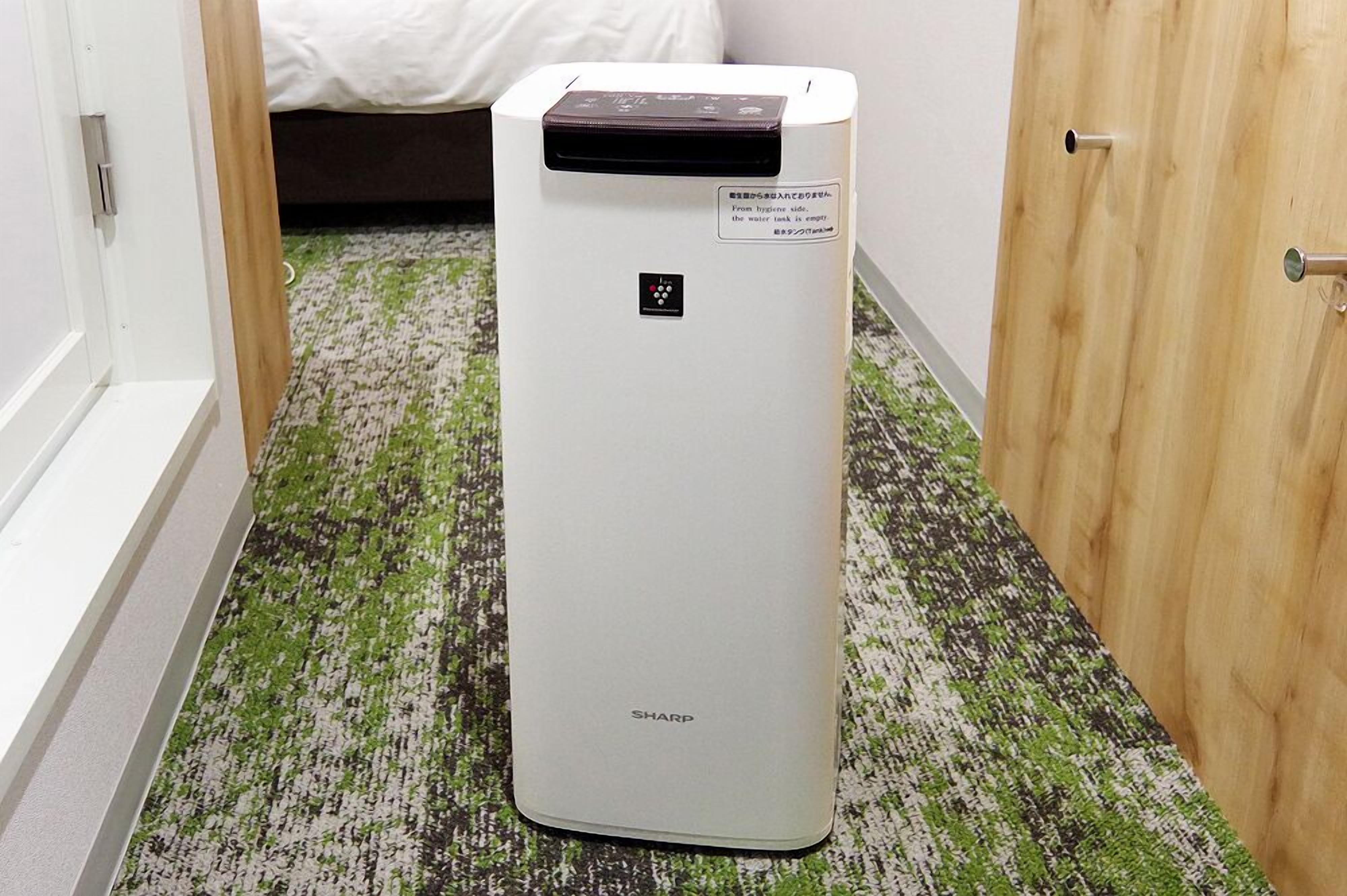 Purificador de aire blanco con panel de control y etiqueta en una habitación con alfombra estampada y una cama al fondo.