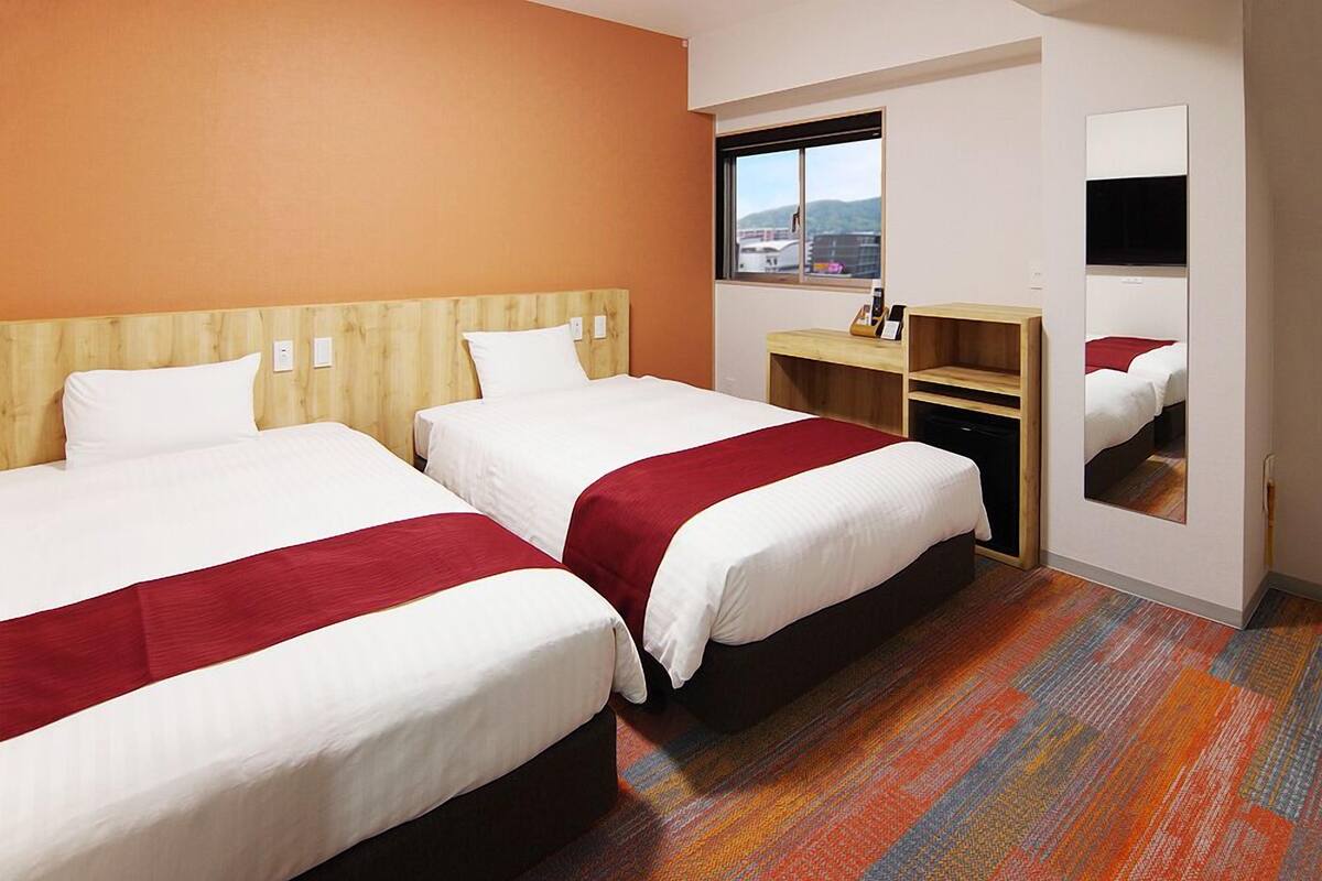 A hotel room with two beds, a wooden headboard, a small desk, a mirror, and a window with a city view.