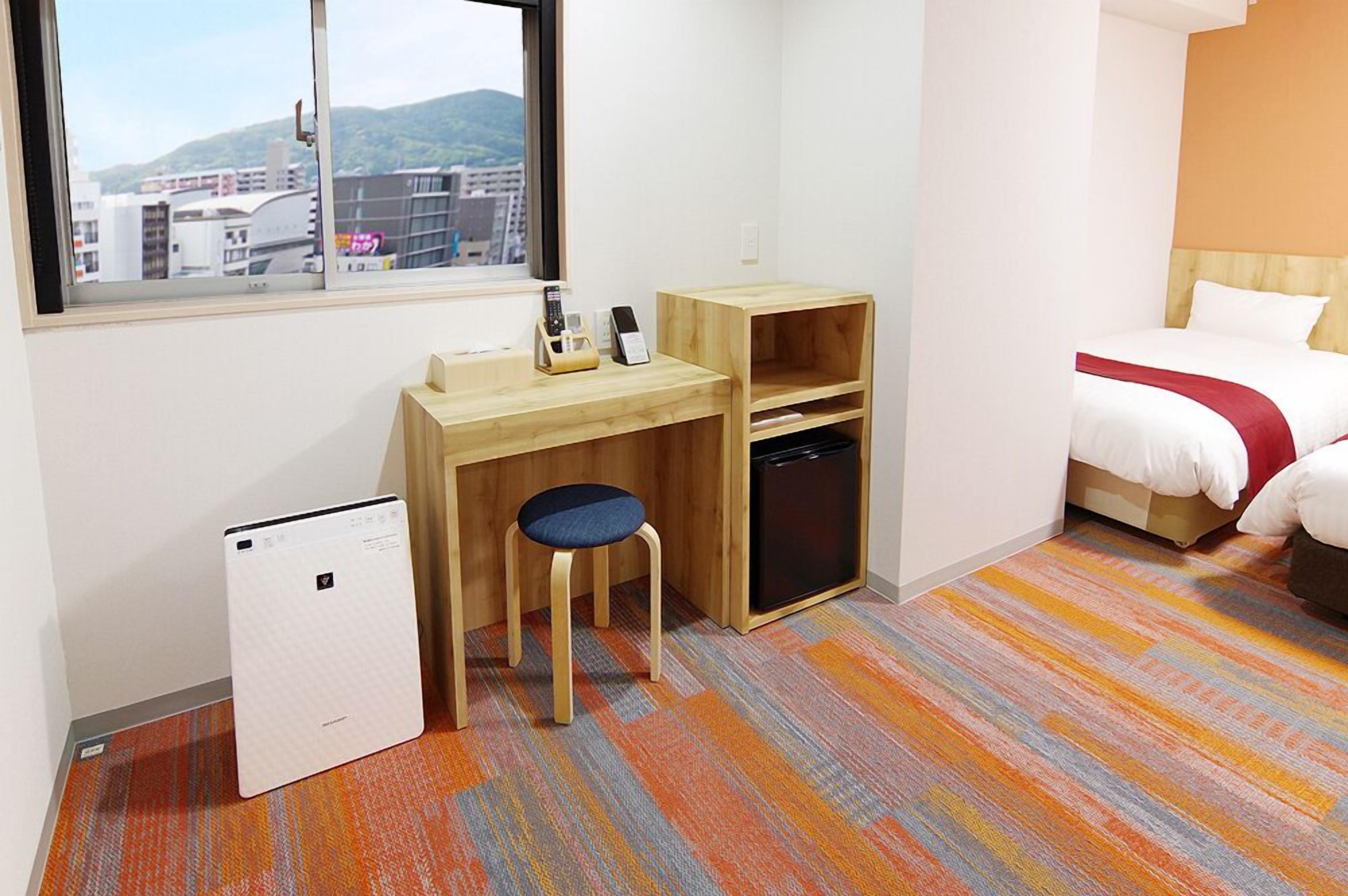A hotel room with a bed, a desk, a chair, a small refrigerator, and a window with a city view.