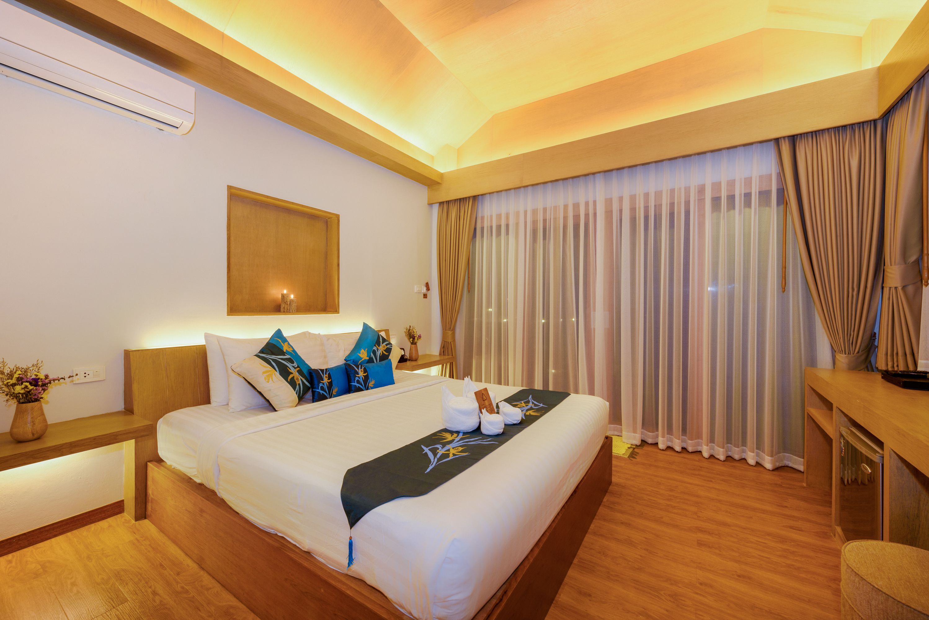A hotel room with a large bed, wooden headboard, bedside tables, a nightstand, a desk, a chair, a window with curtains, and an air conditioning unit.