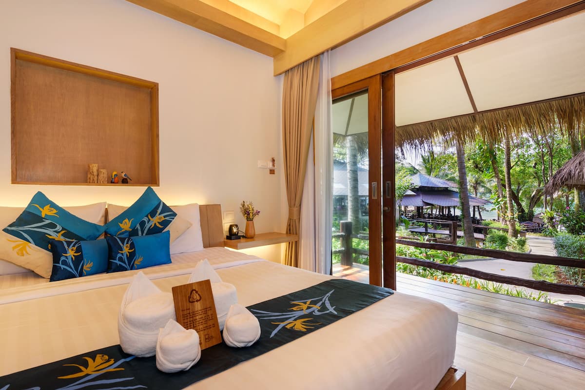 A hotel room with a large bed, a wooden headboard, a view of the garden, and a balcony with umbrellas.