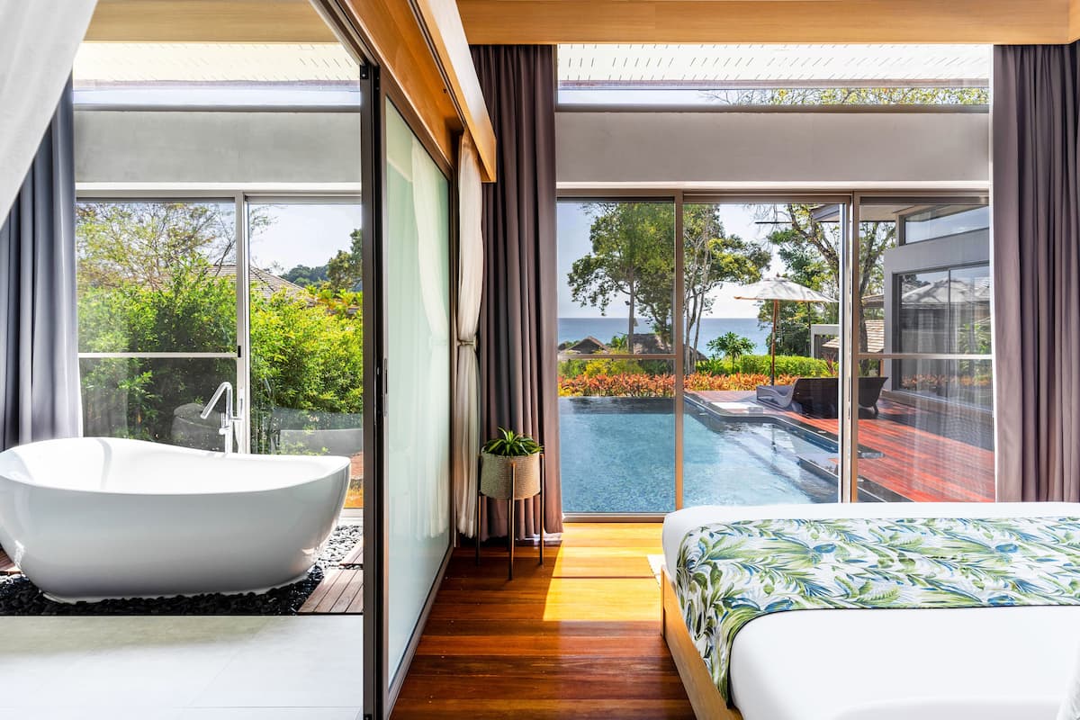 A modern bedroom with a large bathtub, a bed with a floral bedspread, and a view of a swimming pool and garden.